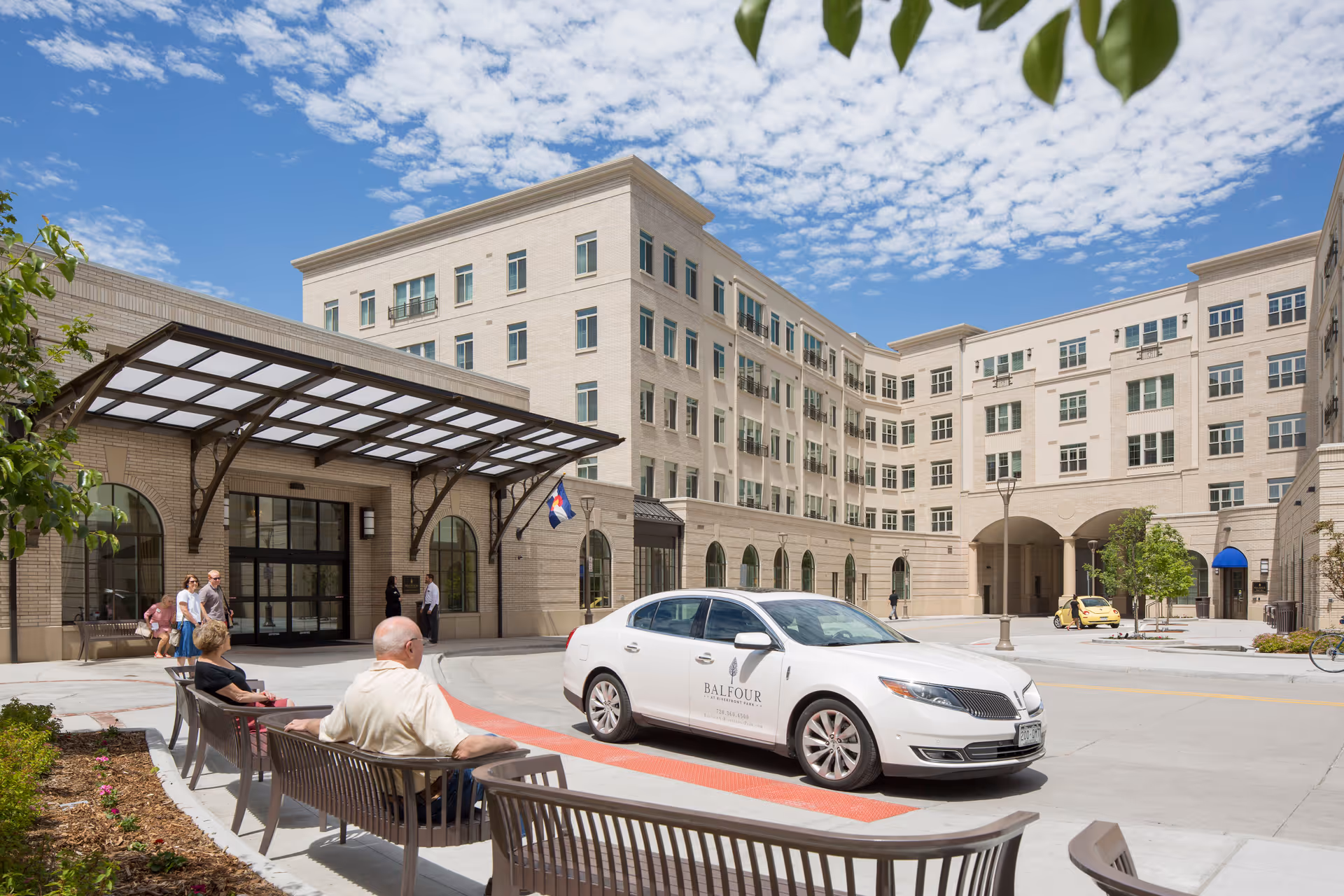 Exterior view of Balfour at Riverfront Park senior living facility on a sunny day with a white car parked in front displaying the Balfour logo. Several people are sitting on benches near the entrance, and others are walking nearby. The building is multi-story with many windows and a covered entrance.