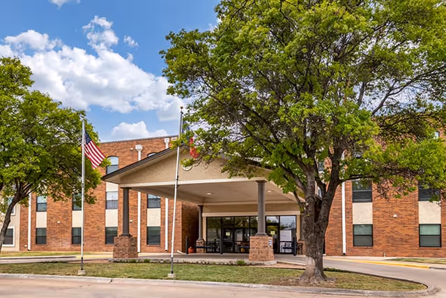 Front entrance of a brick senior living building with a covered porte-cochère, flags, and trees.