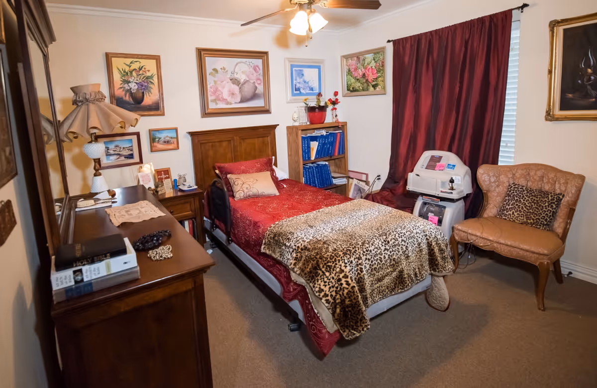 A cozy, warmly decorated bedroom in a senior living facility with a single bed covered by a leopard-print throw, dresser, armchair, and medical equipment by a curtained window.