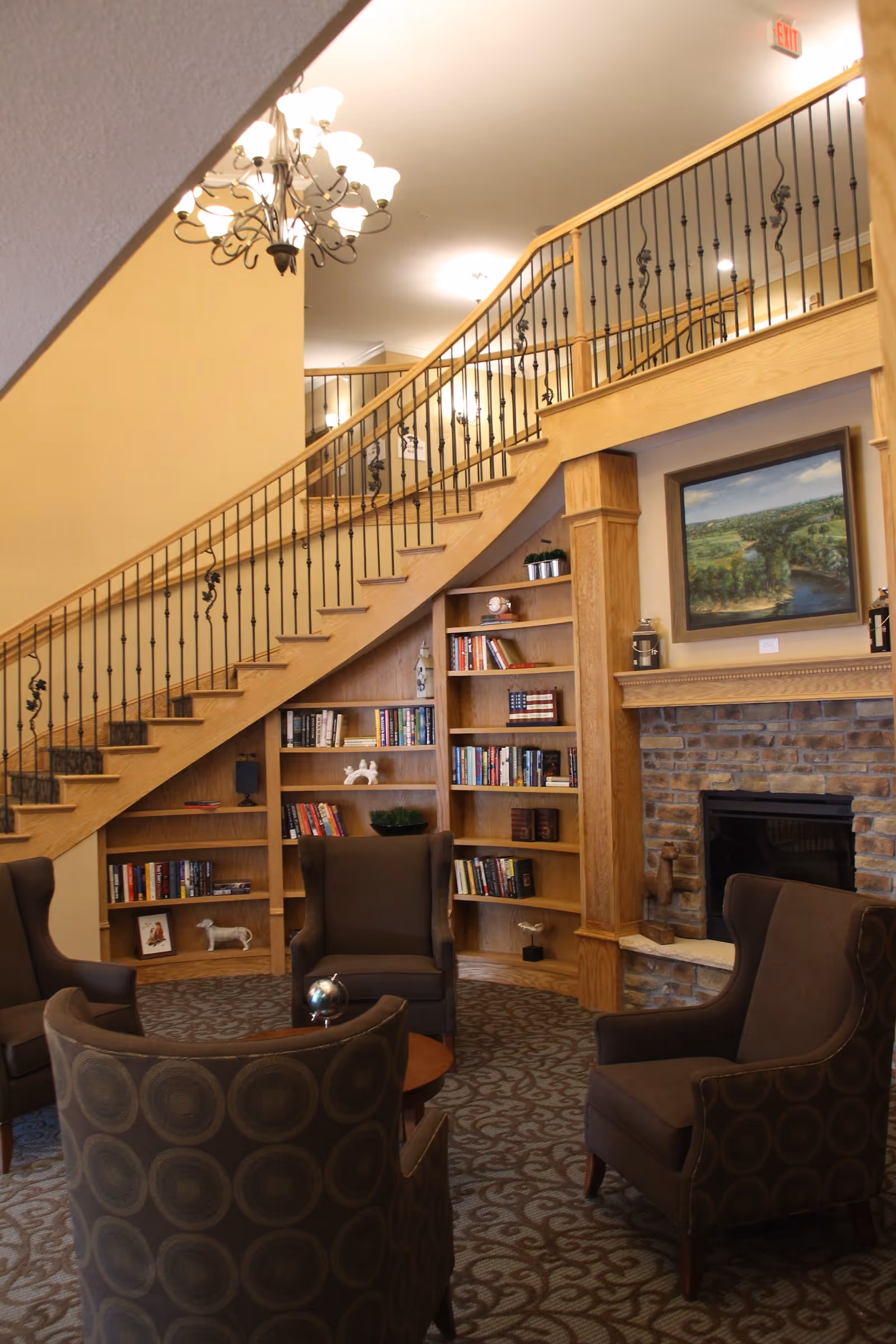 A cozy senior living common area featuring a curved wooden staircase with black wrought iron railings. Below the staircase is a built-in wooden bookshelf filled with books and decorative items. There are four upholstered armchairs arranged around a small wooden table on a patterned carpet. A brick fireplace with a wooden mantle and a landscape painting above it is visible on the right side. Warm lighting from ceiling fixtures and a chandelier brightens the space.