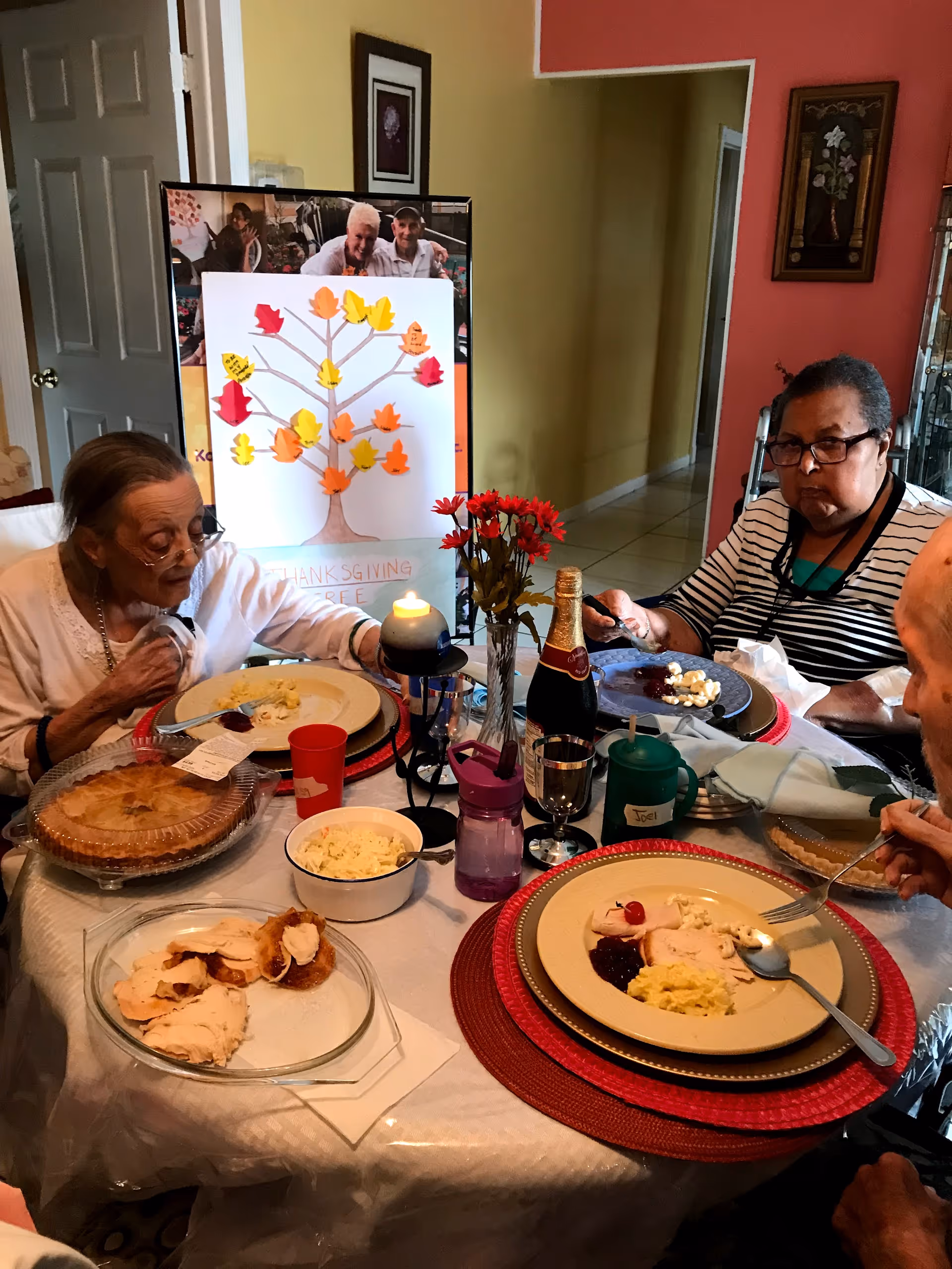Three elderly people sitting around a dining table enjoying a meal with plates of food including turkey, mashed potatoes, and cranberry sauce. The table is decorated with a vase of red flowers, a lit candle, and a bottle of sparkling beverage. In the background, there is a Thanksgiving themed poster with a tree and colorful paper leaves.