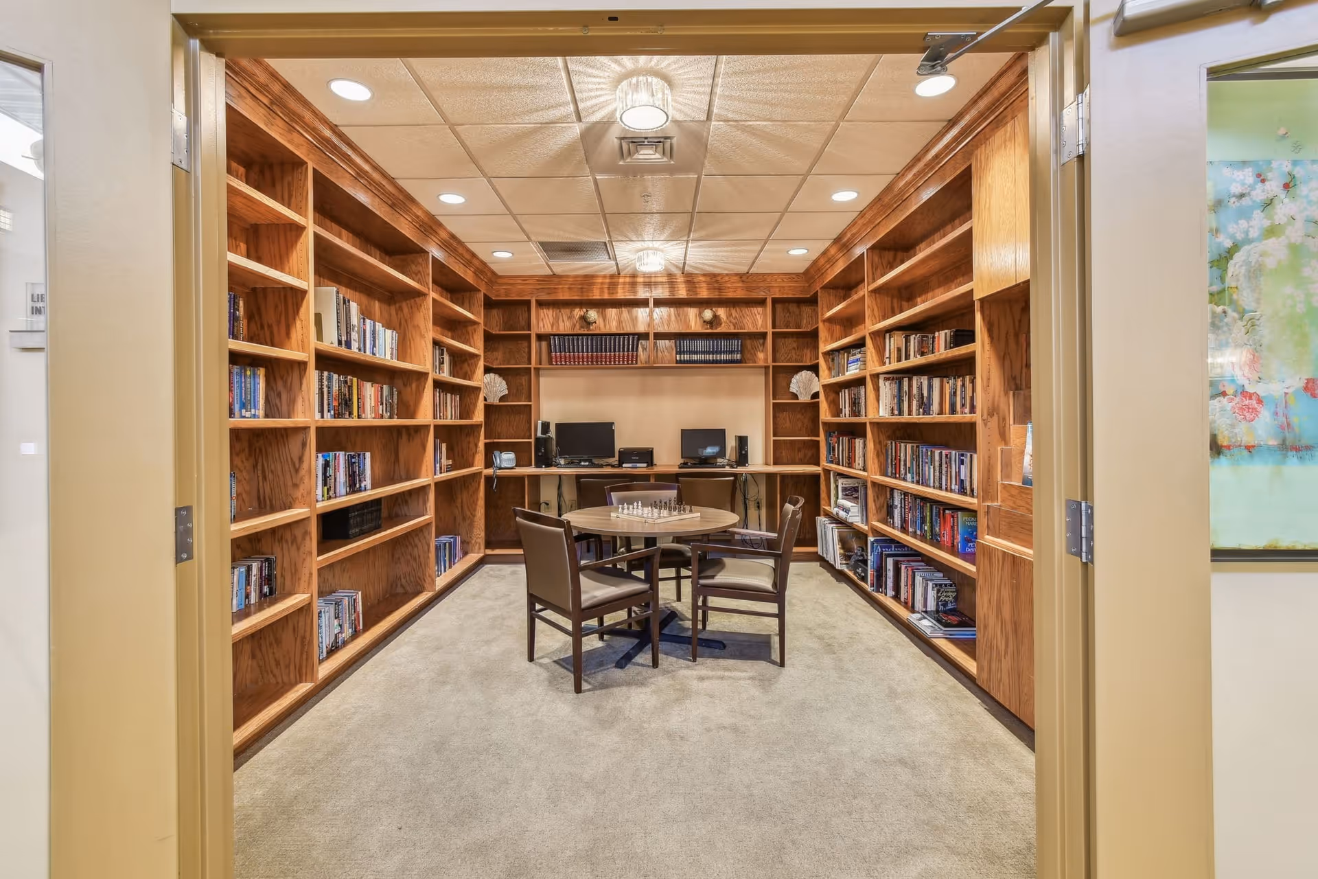 A cozy library room with wooden bookshelves lining three walls filled with books. In the center, there is a round table with four chairs and a chessboard on top. At the back of the room, there are two computer workstations with monitors and speakers. The room has a beige carpet and a ceiling with recessed lighting and two decorative light fixtures.
