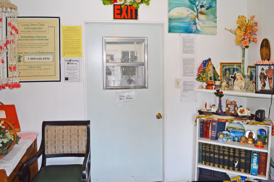 Interior view of a room with a white door that has a small window and an exit sign above it. To the left of the door is a framed notice about Long-Term Care Ombudsmen and a chair with patterned upholstery. To the right of the door is a white shelf filled with books, decorative items, religious statues, and flowers. A colorful painting hangs above the shelf.