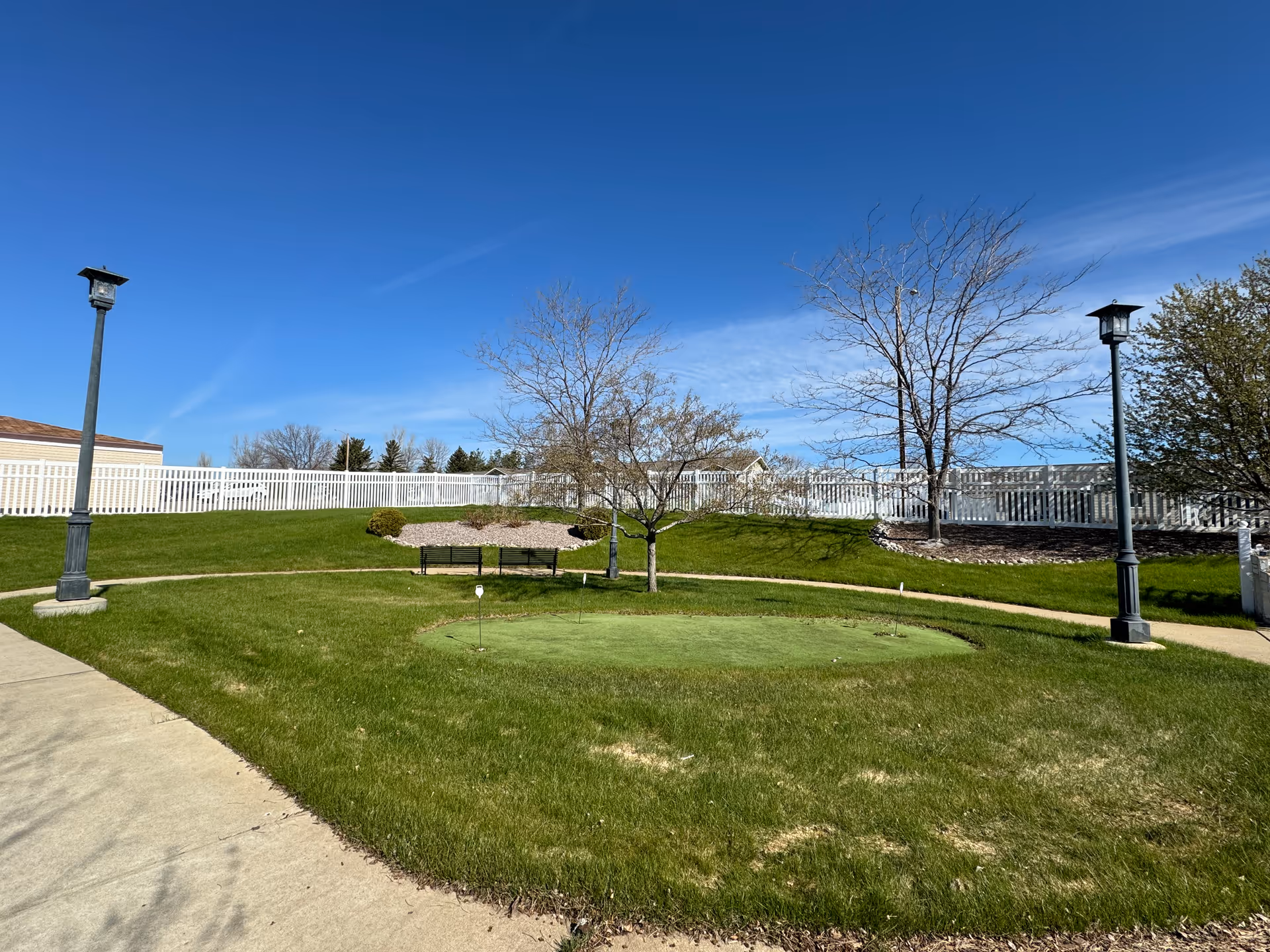 Well-kept outdoor lawn with a circular putting green, benches, lamps, and white fencing.