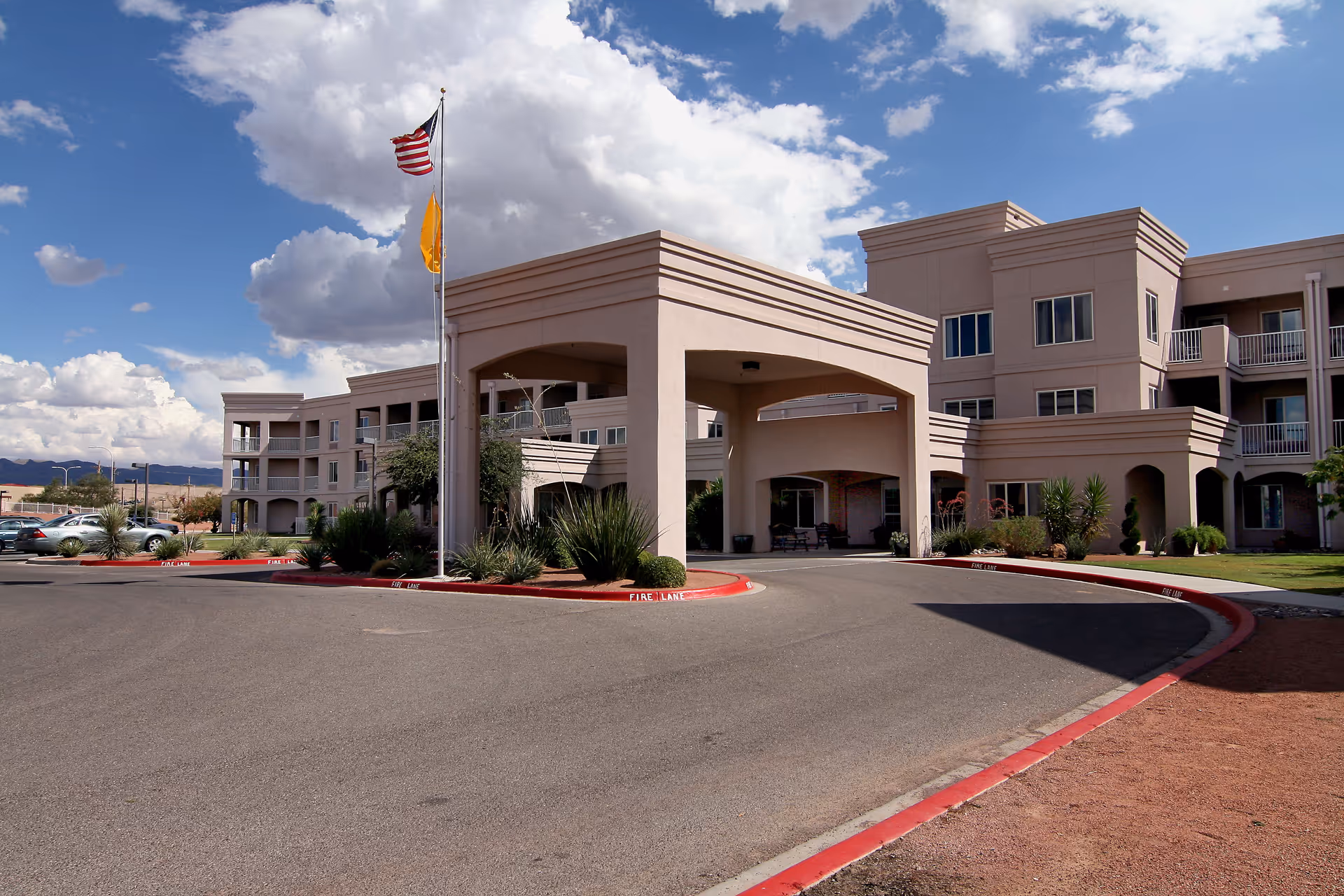 Exterior view of Solstice Senior Living at Las Cruces showing a large beige building with multiple windows and balconies, a covered entrance with columns, two flagpoles with the American flag and another flag, surrounded by landscaping and a parking area under a partly cloudy sky.