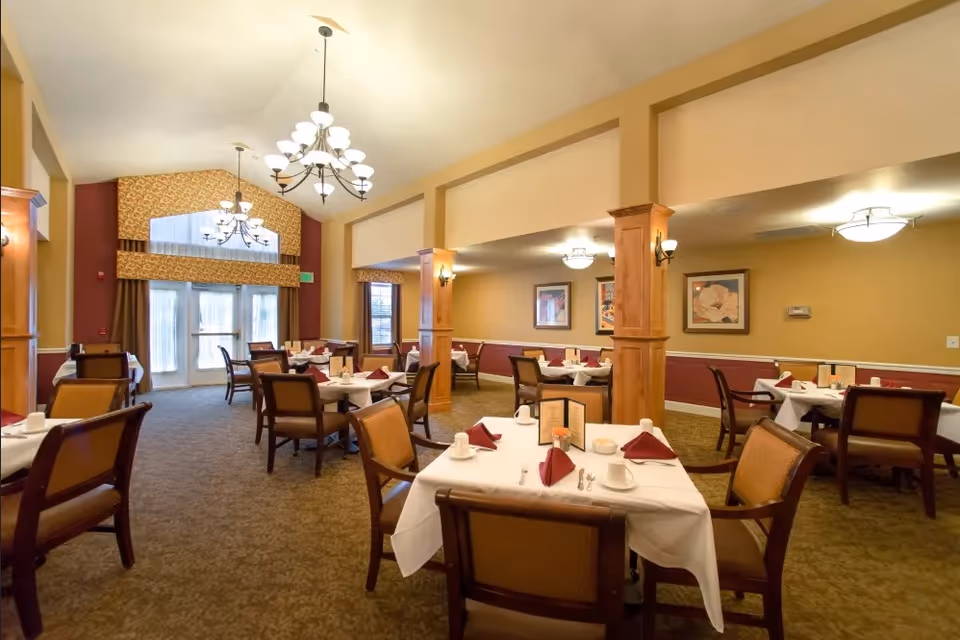 Spacious, well-lit dining room with multiple tables set with white linens, folded napkins, chandeliers, and upholstered chairs.