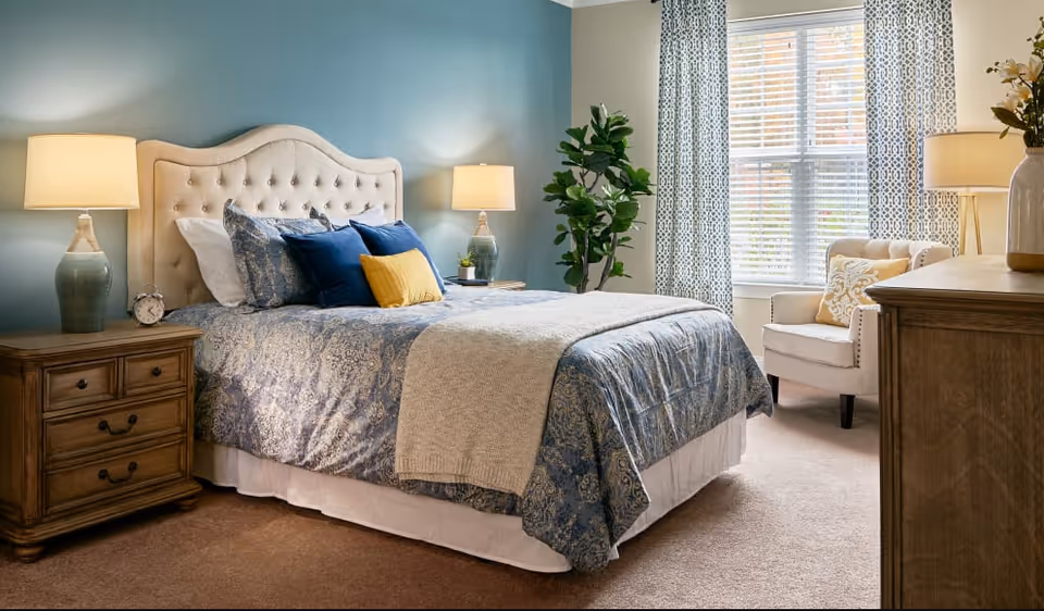 A furnished bedroom with a tufted headboard, patterned bedding, bedside tables with lamps, an armchair by the window, and a potted plant.