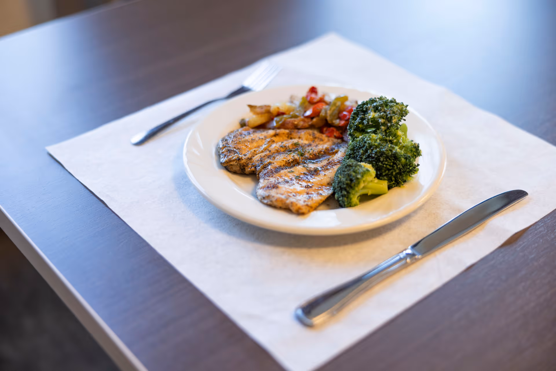 A plate with grilled chicken, broccoli, and roasted vegetables on a placemat with a fork and knife on a table.