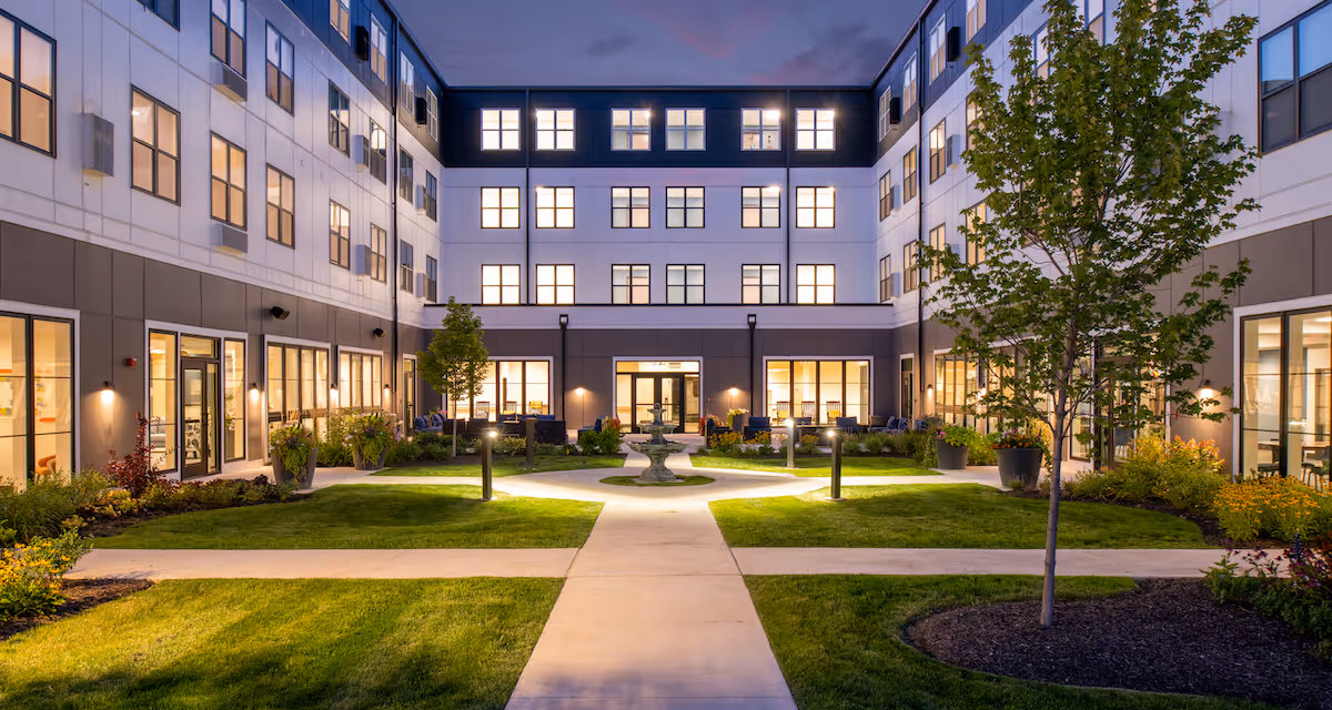 Well-lit central courtyard of a multi-story senior living building at dusk with walkways, lawn, trees, and windows.