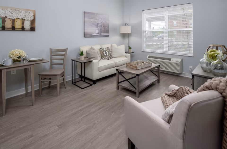 A cozy living room with a beige armchair draped with a knitted blanket, a cream-colored sofa with decorative pillows, a wooden coffee table with a tray, a small dining table with two chairs, and a window with blinds letting in natural light. The room has light gray walls, wood flooring, and decorative wall art and plants.