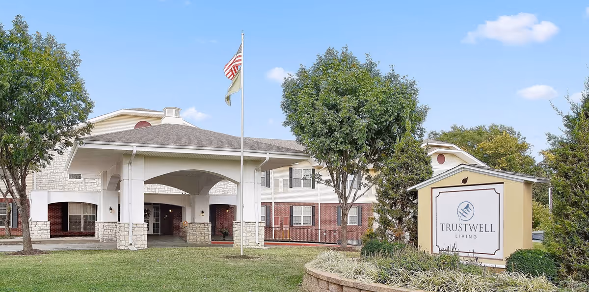 Exterior view of Trustwell Living of Raytown facility showing the main entrance with a covered driveway, two flagpoles with flags, trees, and a sign with the facility name on a landscaped area.