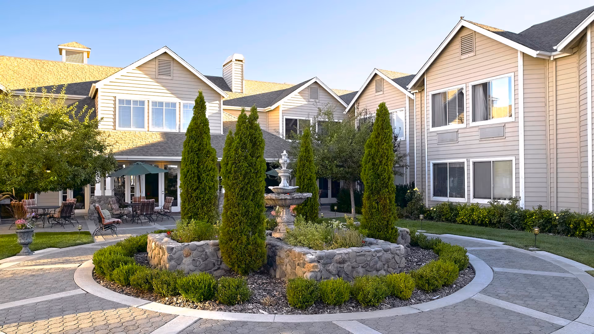 Outdoor courtyard area at a senior living facility with a circular stone planter containing tall evergreen shrubs and a multi-tiered water fountain in the center. Surrounding the planter is a paved walkway and patio seating with tables and chairs under umbrellas. The background shows beige two-story buildings with multiple windows and a clear sky.