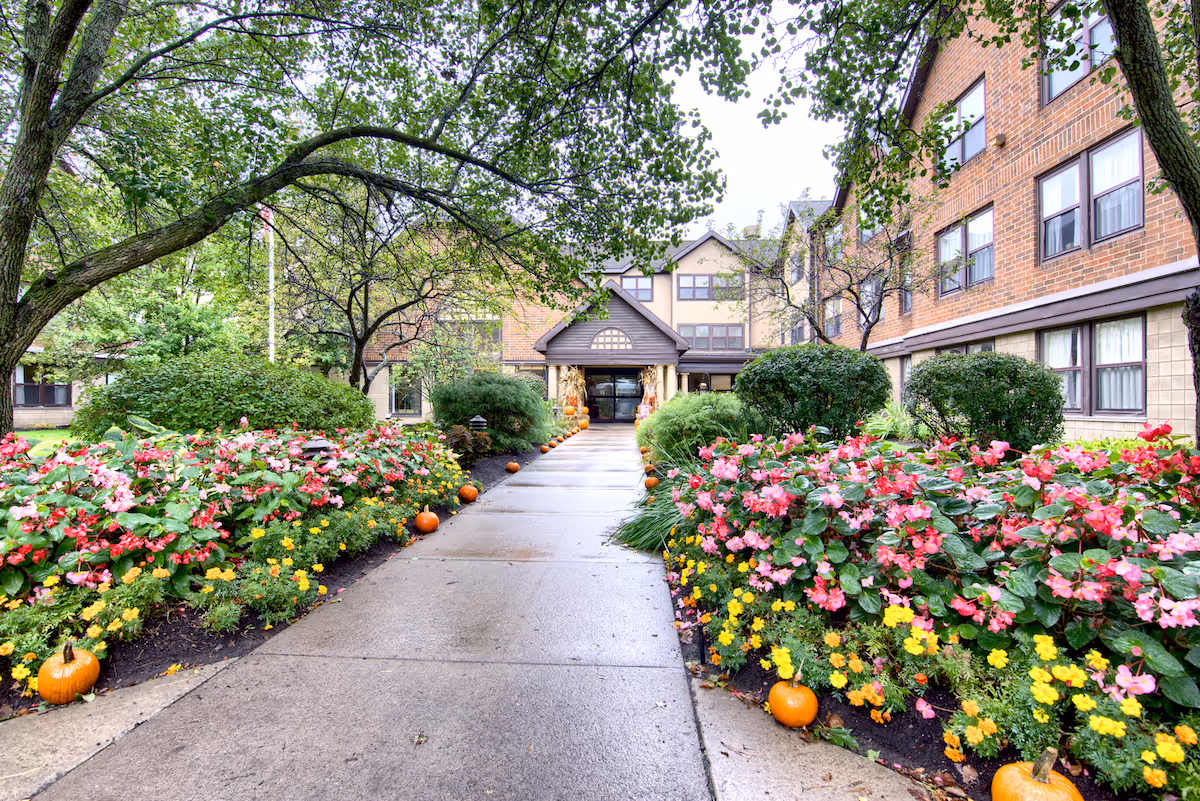 A paved walkway lined with small pumpkins and colorful flower beds leads to the entrance of a multi-story brick senior living facility building. Trees and shrubs surround the path, and the building has multiple windows and a covered entryway.