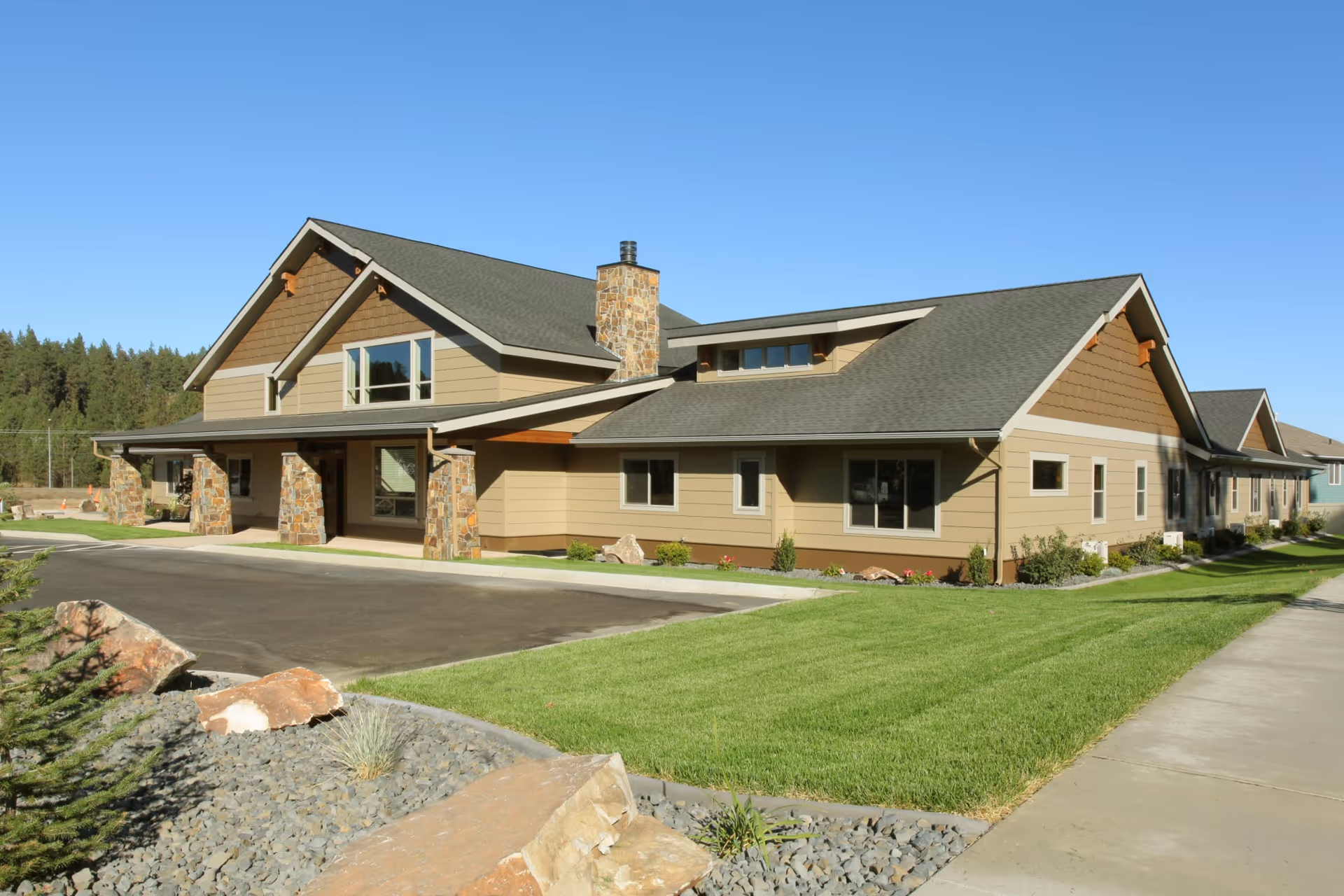 Exterior view of The Lodge at Riverside Harbor, a single-story building with a stone chimney and stone pillars supporting a covered entrance. The building has beige siding with brown accents and a gray shingled roof. The surrounding area includes a paved driveway, green lawn, rocks, and a sidewalk under a clear blue sky.