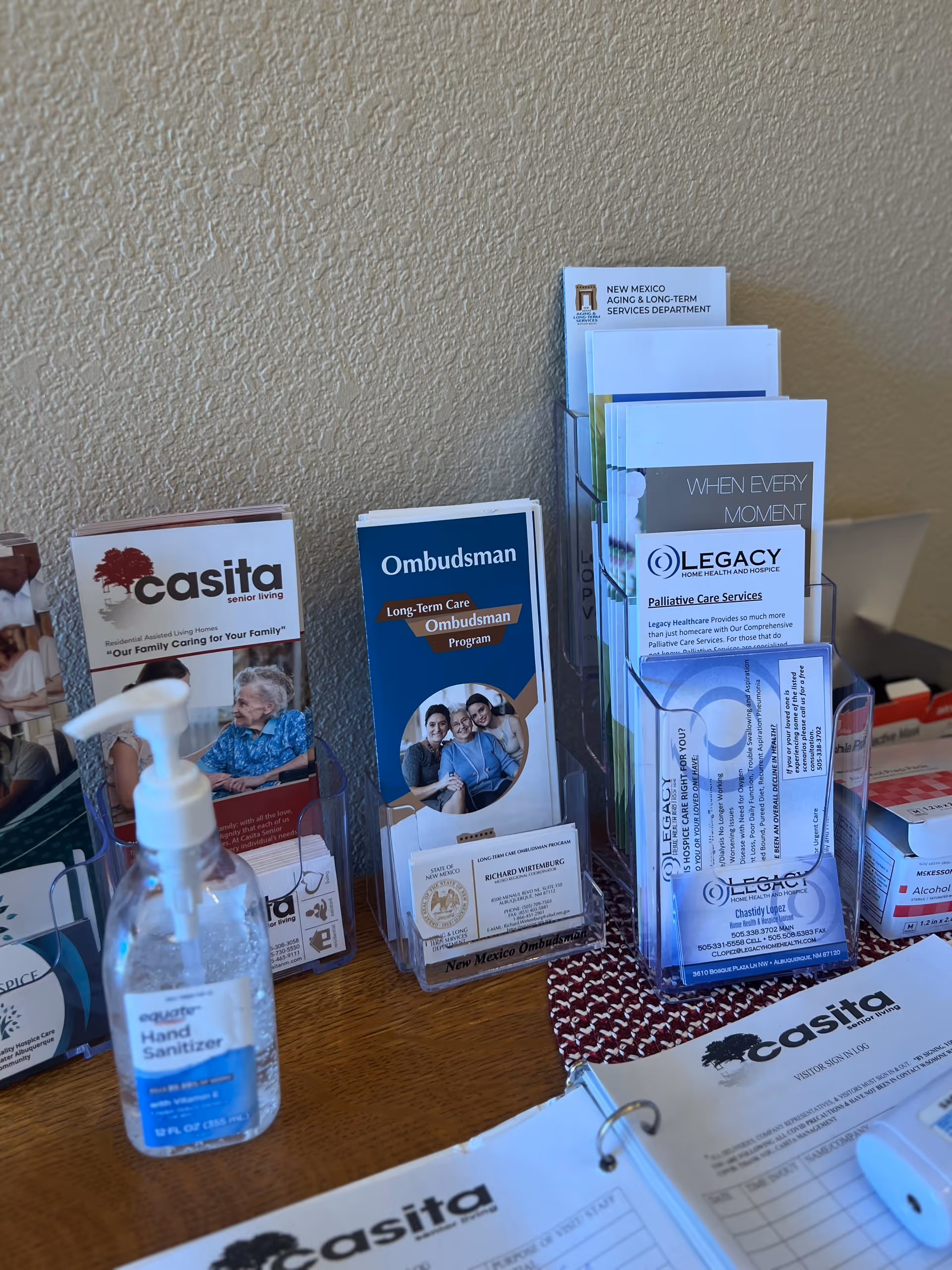 A wooden table with various informational brochures and pamphlets displayed in clear holders. Visible brochures include Casita Senior Living, Ombudsman Long-Term Care Program, and Legacy Home Health and Hospice. A bottle of hand sanitizer is also on the table, along with sign-in sheets labeled Casita Senior Living.