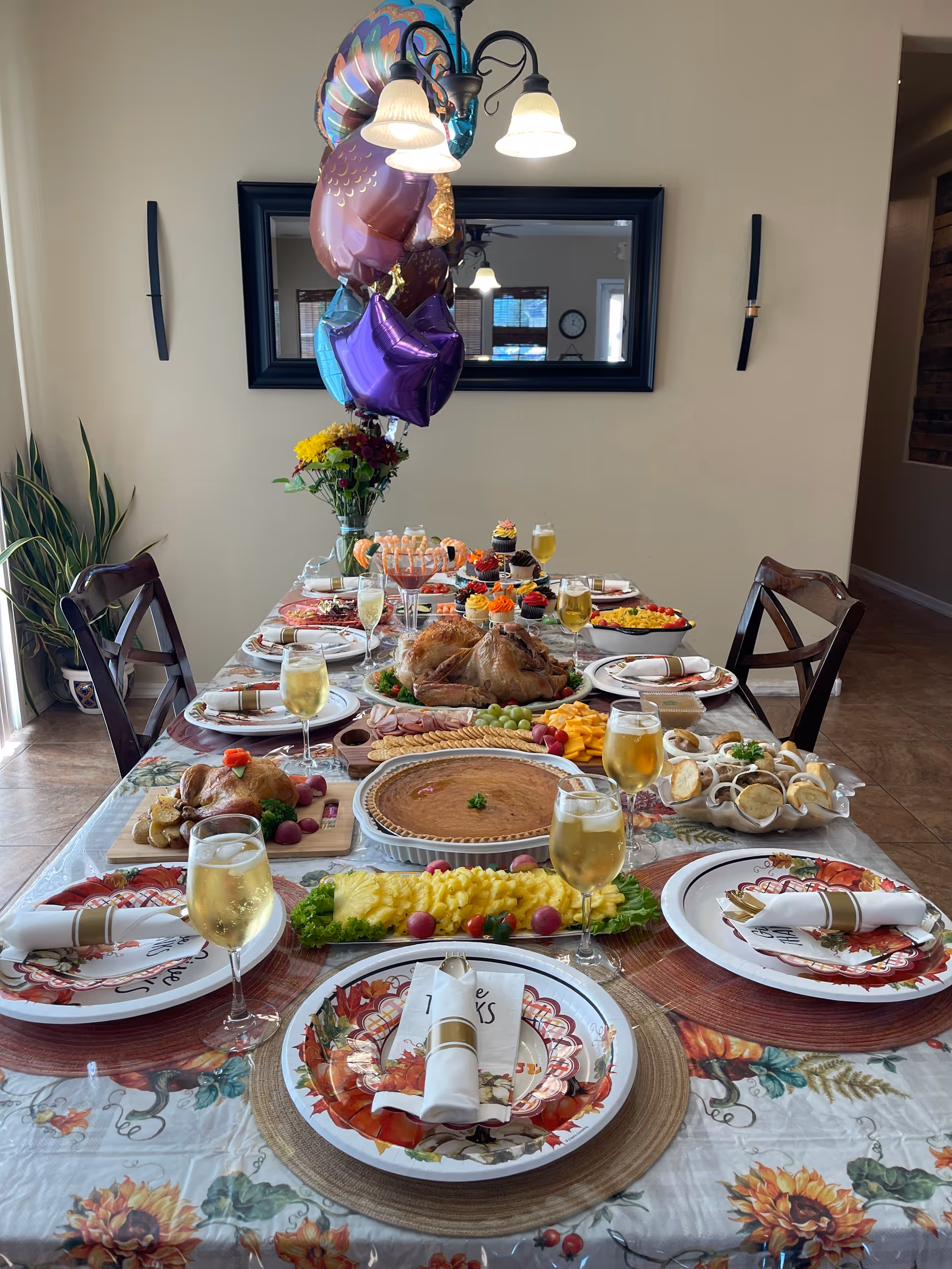 A dining table set for a festive meal with plates, napkins, and glasses filled with a light beverage. The table is decorated with a floral tablecloth and features a variety of foods including a roasted turkey, pie, cheese, grapes, cupcakes, and other dishes. There are colorful balloons tied to a vase with flowers at the center of the table. The room has wooden chairs and a large mirror on the wall reflecting a clock and window.