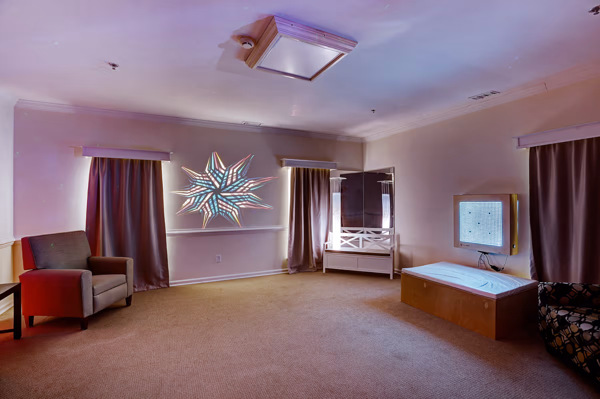 A spacious room with beige carpet and light-colored walls featuring two windows with closed curtains. The room contains a gray armchair on the left, a white bench with a mirror in the corner, and a wooden box with a light panel mounted on the wall above it. A colorful star-shaped light decoration is projected on the wall between the windows.