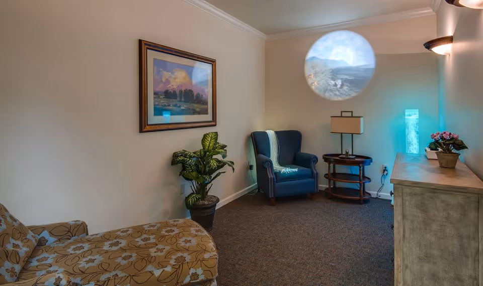 A cozy living room area with a floral patterned chaise lounge on the left, a potted plant next to it, and a framed landscape painting on the wall. On the right side, there is a blue armchair with a light blue throw draped over it, a round wooden side table with a lamp, and a wooden cabinet with a small potted plant and a tall illuminated decorative piece emitting blue light. The walls are light beige, and the carpet is dark brown.