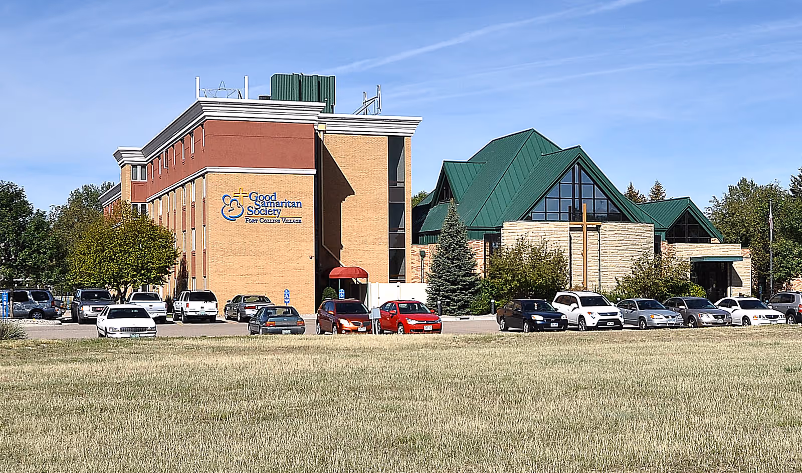 Exterior view of Good Samaritan Society - Fort Collins Village building with a parking lot in front and several cars parked. The building has a combination of brick and stone walls with a green roof and a large cross mounted on the stone section. Trees and a grassy area are visible around the building under a clear blue sky.