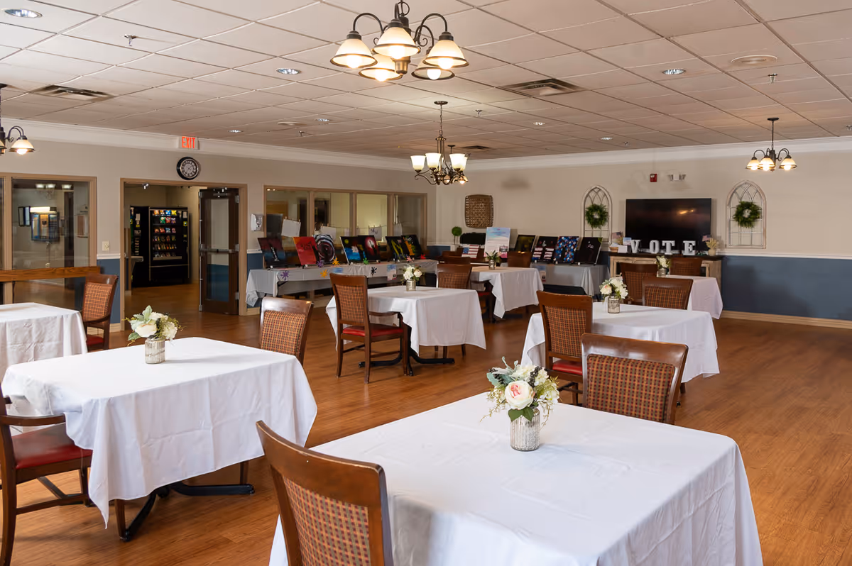 Spacious dining/activity room with multiple white-clothed tables and chairs, floral centerpieces, and a TV/display at the back.