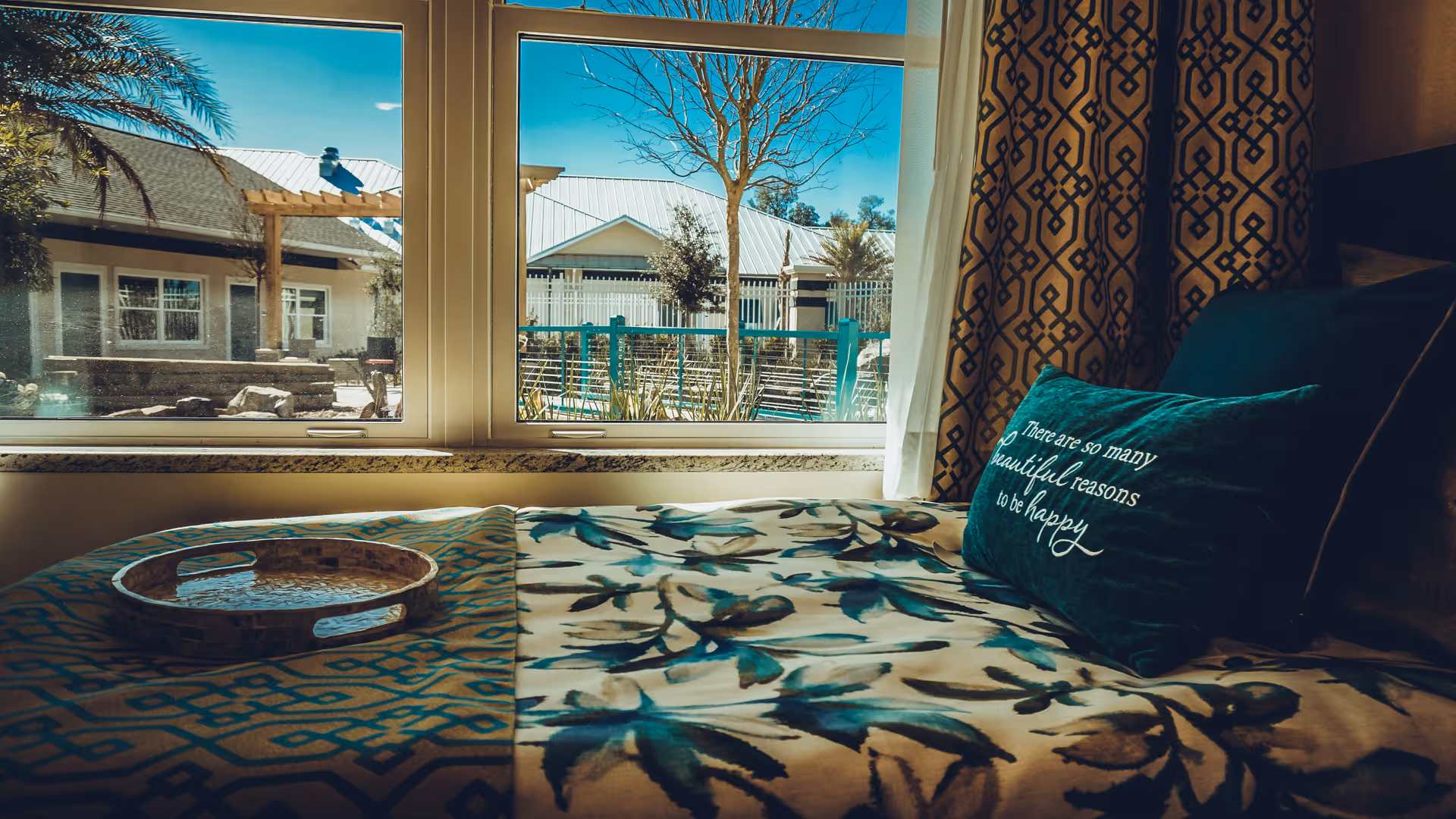 Sunlit bedroom with a patterned bedspread, decorative throw pillow on the bed, and a large window overlooking a courtyard.
