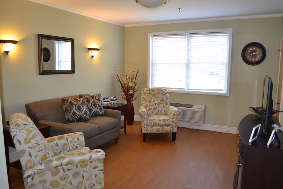 Bright assisted-living sitting room with a sofa, two patterned armchairs, a TV console, wall mirror and a large window.