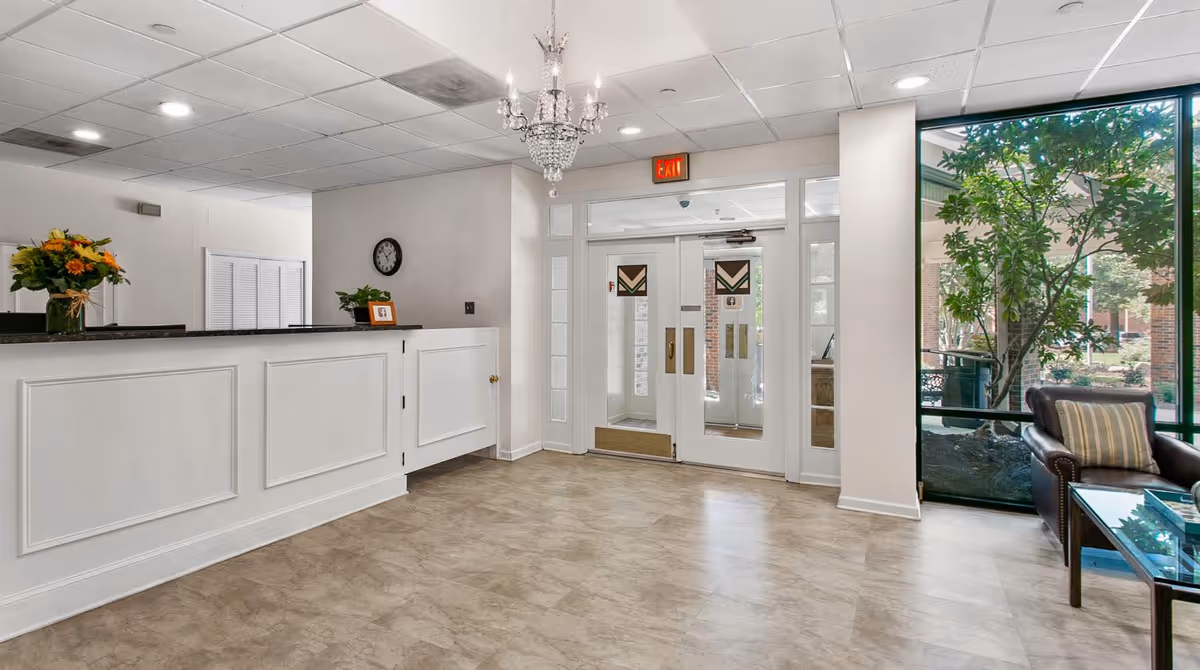 A bright and clean reception area with a white front desk adorned with a vase of flowers and a small framed picture. There is a clock on the wall behind the desk and a chandelier hanging from the ceiling. Double glass doors lead outside, and large windows on the right reveal greenery and outdoor seating. A brown leather armchair with a striped pillow and a glass coffee table are visible near the windows.