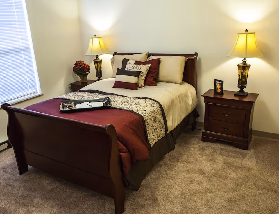 A neatly made bed with a wooden frame in a bedroom. The bed has beige and burgundy bedding with decorative pillows. There are two wooden nightstands on either side of the bed, each with a lamp. One nightstand also has a small framed photo, and the other has a vase with flowers. A window with closed blinds is visible on the left side of the room.