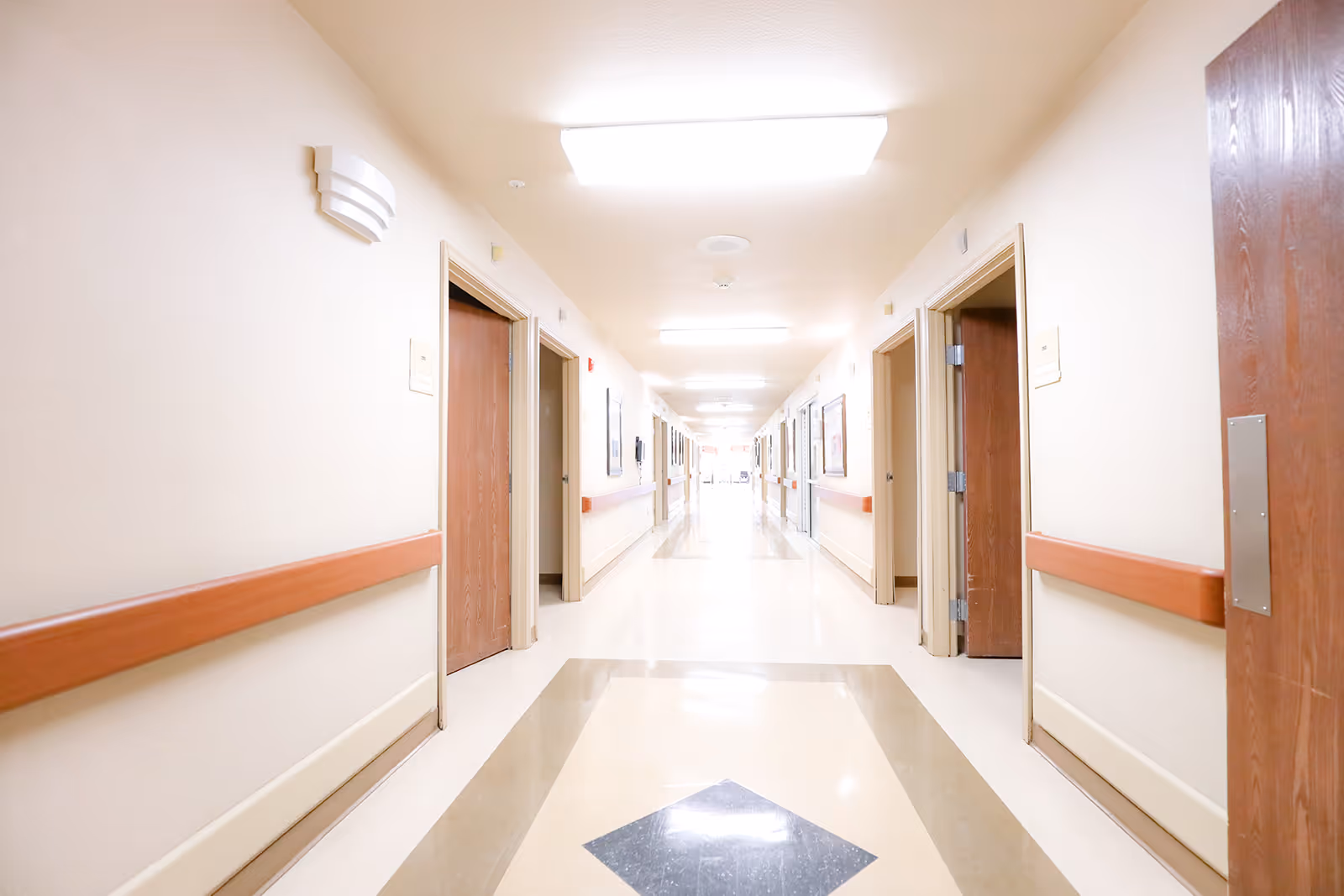 A long, brightly lit hallway in a healthcare or rehabilitation facility with beige walls, wooden doors on both sides, handrails along the walls, and a patterned floor.