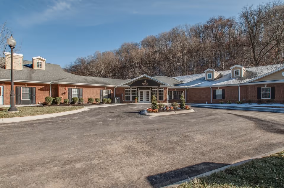 Front exterior view of Barton House Memory Care facility, a single-story brick building with multiple windows, a central entrance with a covered porch, surrounded by a paved driveway and some landscaping. Trees without leaves are visible in the background under a clear blue sky.