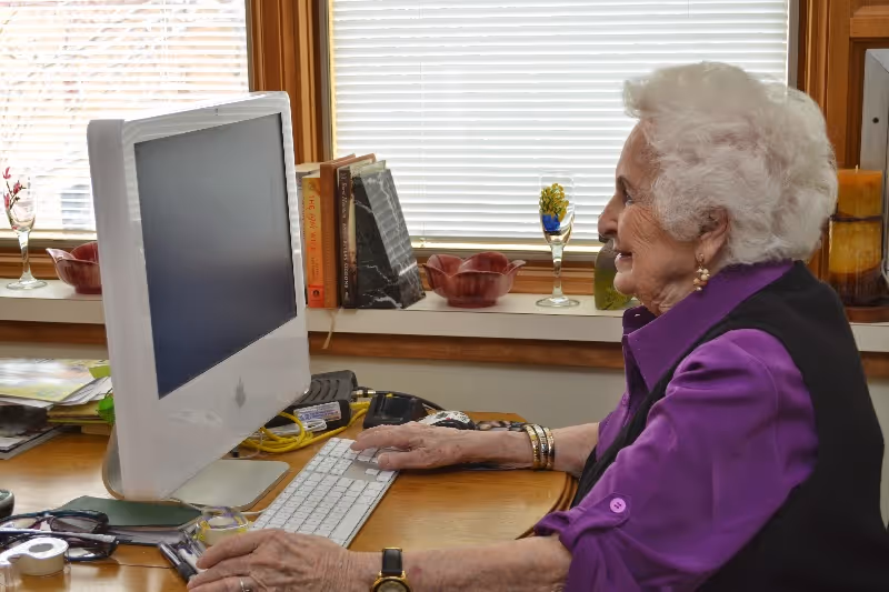 An elderly woman with white hair and wearing a purple shirt and black vest is sitting at a wooden desk using a white iMac computer. The desk has various items including books, glasses, and decorative glassware on the windowsill behind her. The window blinds are partially closed.