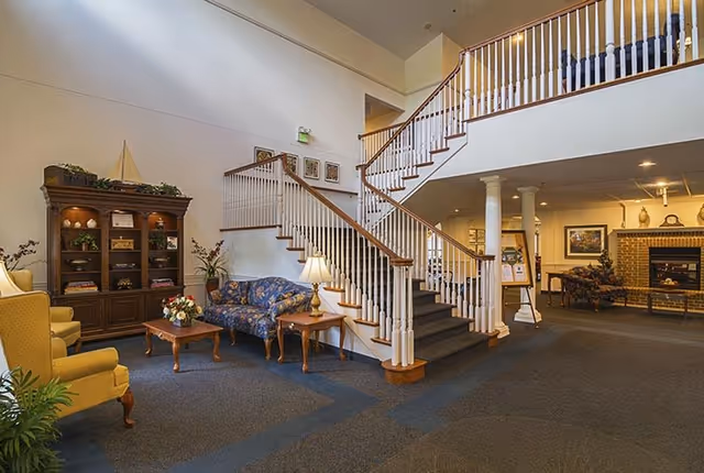 Spacious senior living facility lobby with a central staircase, seating area, cabinet and fireplace in the background.