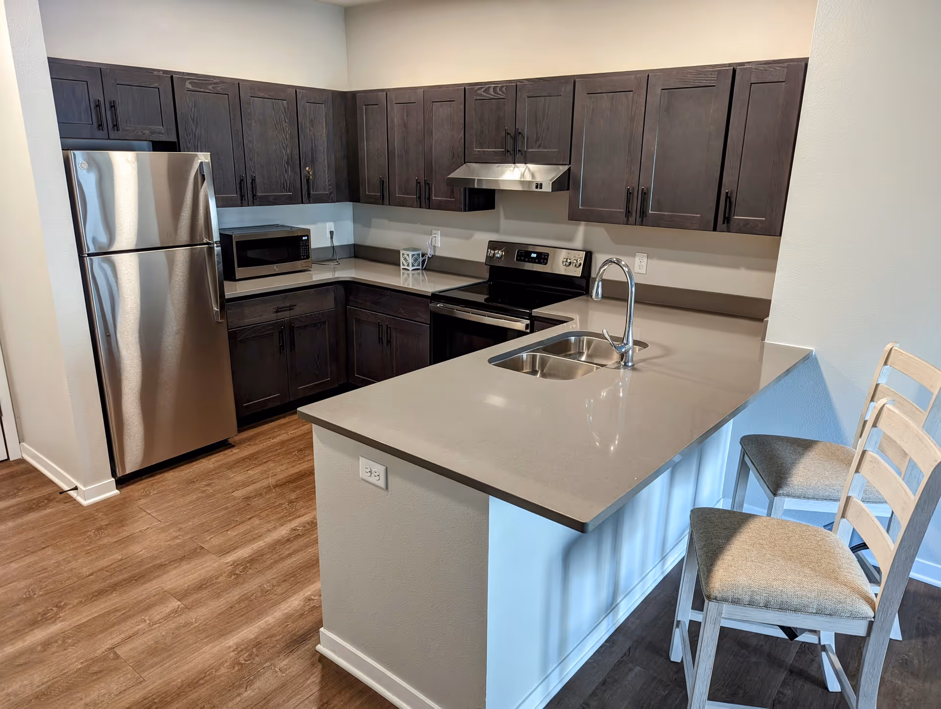 Modern kitchen with dark wood cabinets, stainless steel refrigerator, microwave, stove with range hood, and a countertop with a built-in sink and two cushioned chairs.