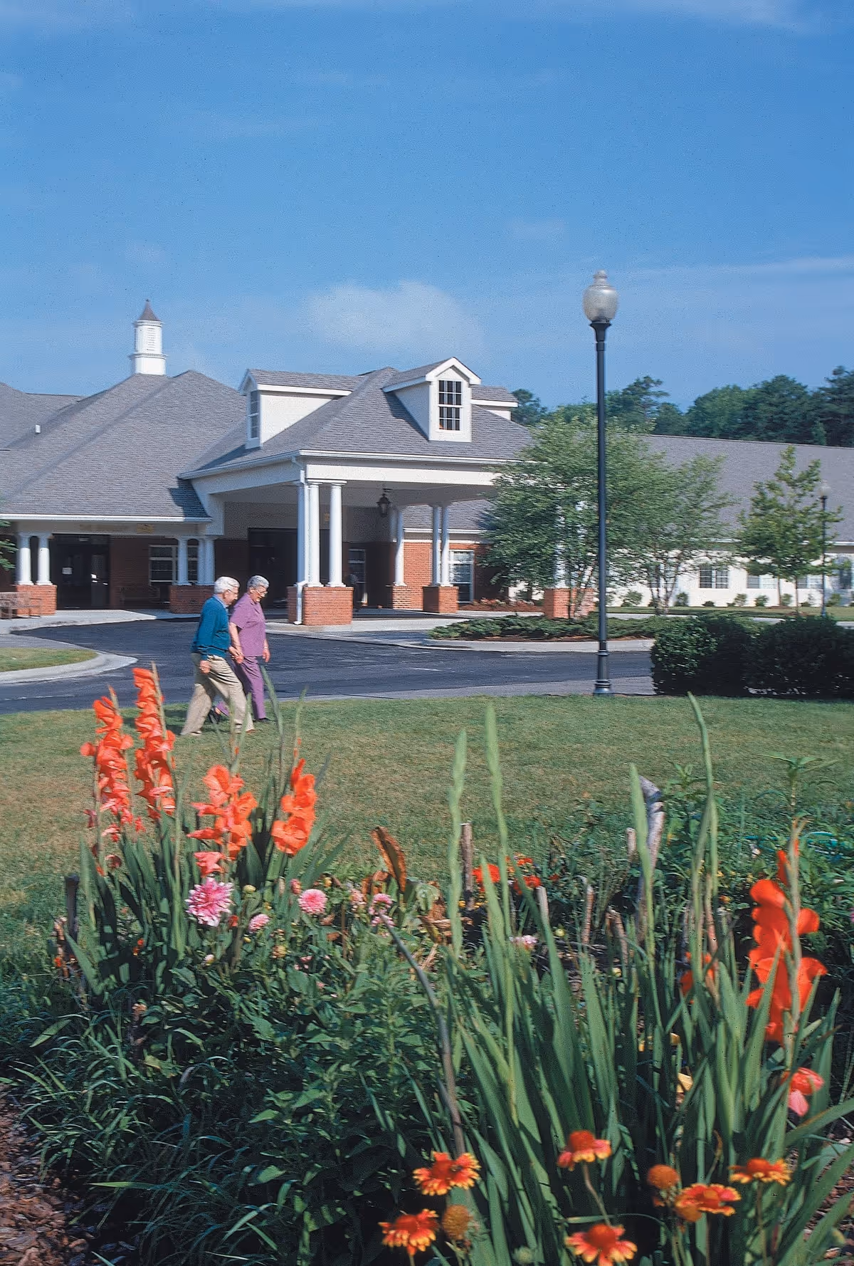 Two elderly people walking on a driveway in front of a senior living facility entrance with flowering beds in the foreground.