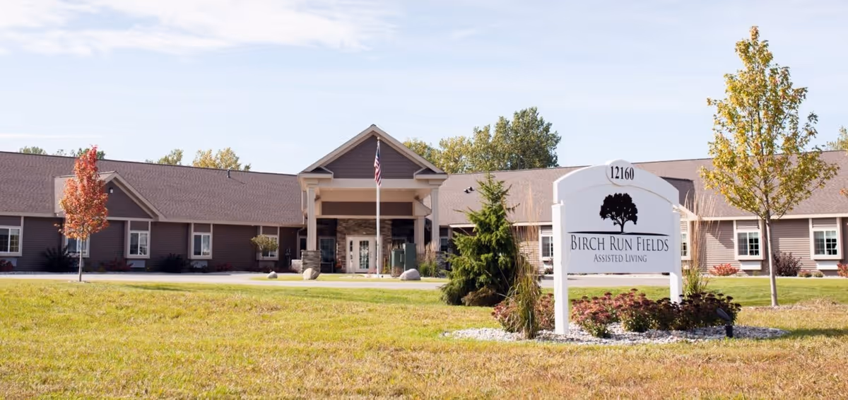 Exterior view of Birch Run Fields Assisted Living facility showing a single-story building with a peaked entrance, an American flag in front, and a landscaped lawn with trees and shrubs. A white sign with the facility's name and address is prominently displayed in the foreground.
