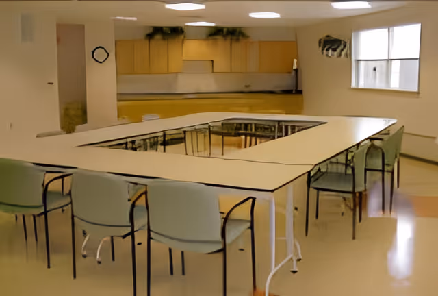 Large institutional dining/activity room with tables arranged in a U-shape and chairs around them, pale walls and windows.