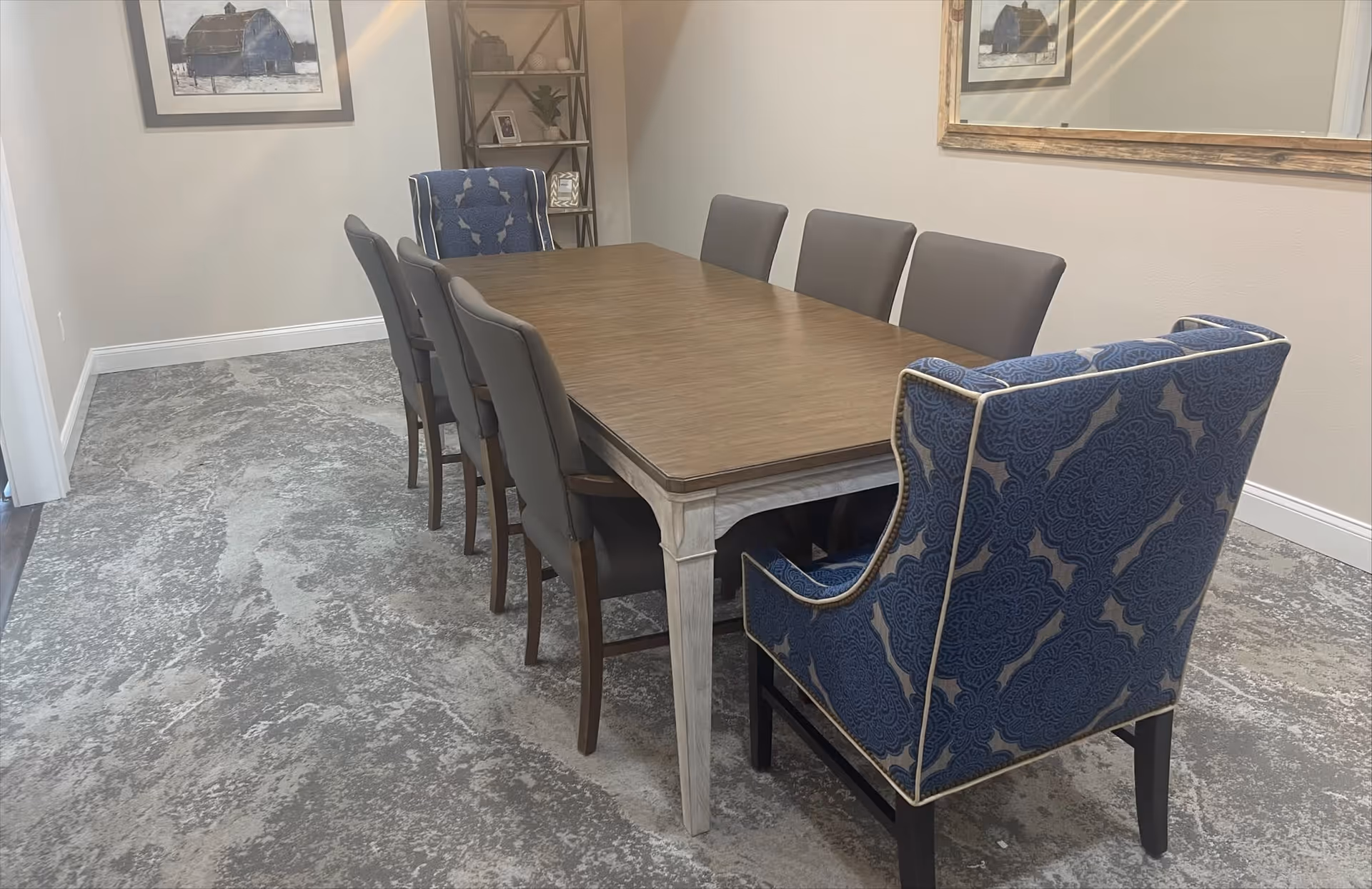 A dining room with a long wooden table surrounded by eight chairs, two of which have blue patterned upholstery with white trim. The room has light-colored walls, a large mirror on one wall, a framed picture of a barn on another wall, and a corner shelf with decorative items.