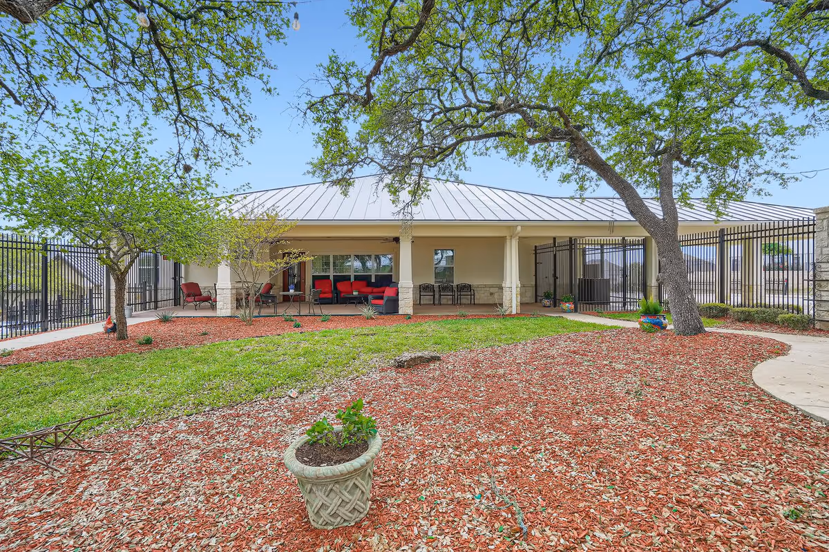 Outdoor garden area with a covered patio featuring red cushioned seating and black chairs. The garden has green grass, mulch, a few small trees, and potted plants. A black metal fence surrounds the area under a clear blue sky.