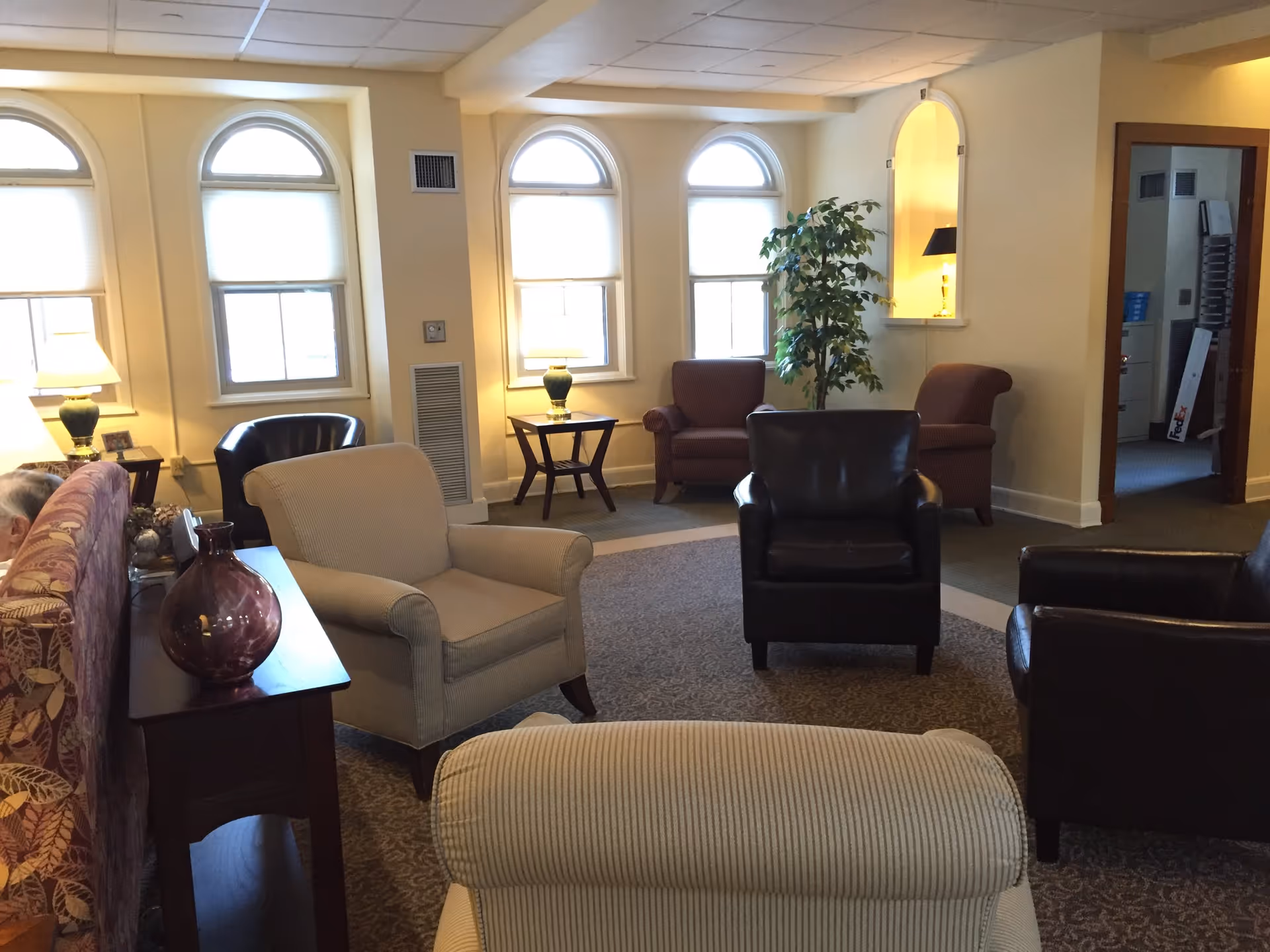 A cozy senior living facility common area with multiple armchairs arranged in a circle on a carpeted floor. The room has three arched windows with blinds, two table lamps on side tables, a potted plant, and a decorative niche with a lamp. The walls are painted light yellow, and there is an open doorway leading to another room.