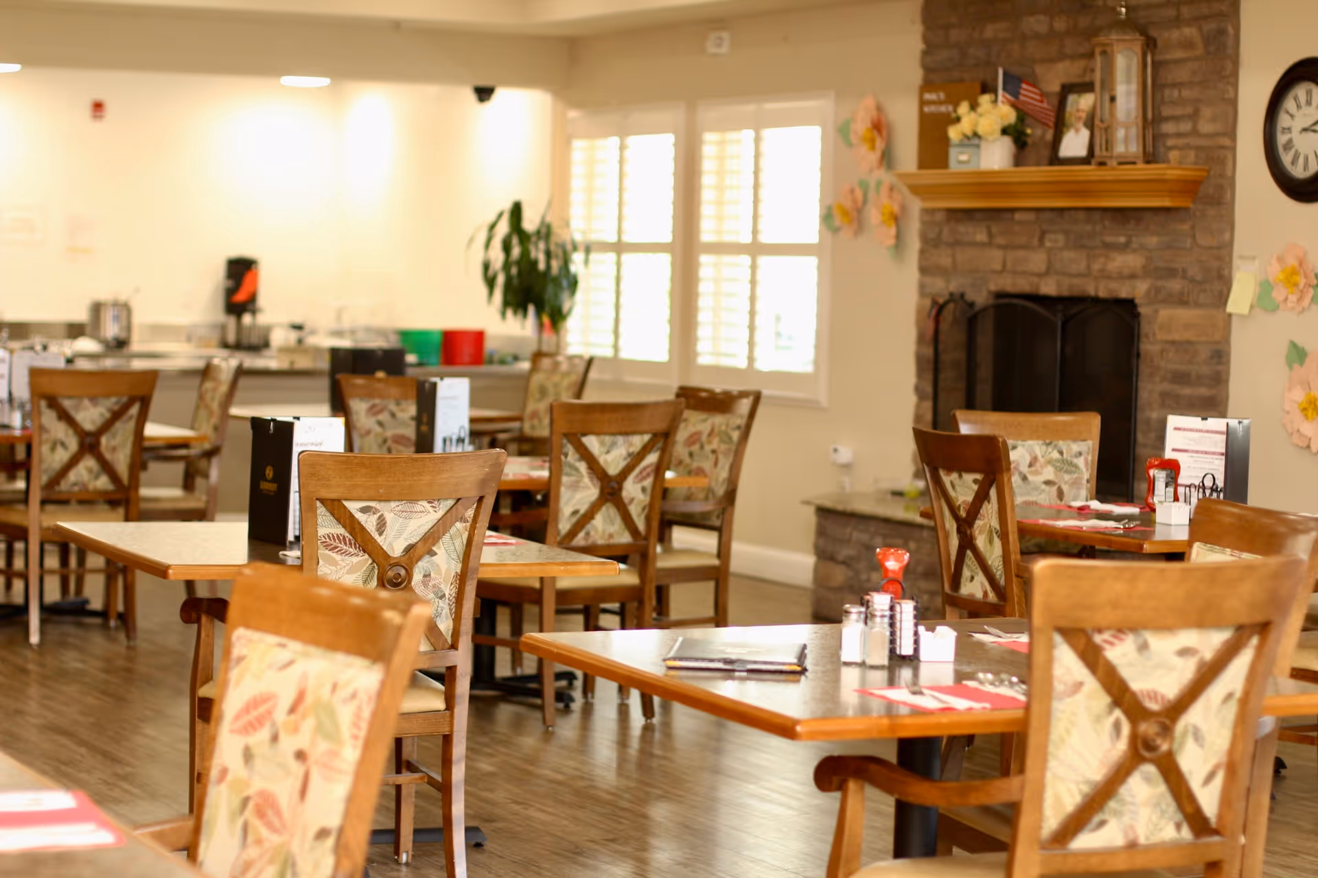 Bright dining room with wooden tables and patterned chairs set for meals, a fireplace, and large windows.