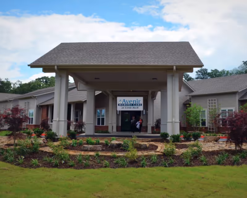 Front exterior view of a single-story memory care facility building with a covered entrance supported by columns, landscaped garden with shrubs and small trees in front, and a sign that reads 'Avenir Memory Care at Little Rock'.