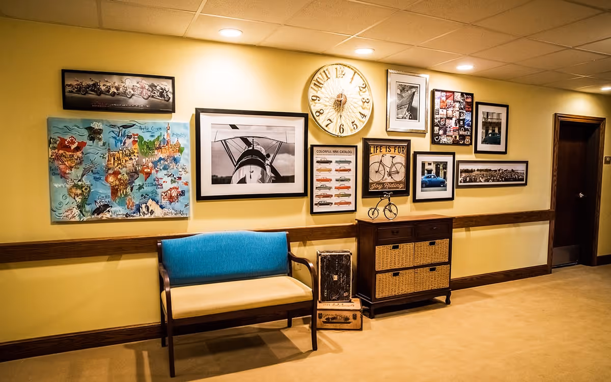A hallway with yellow walls decorated with various framed pictures and a large clock. There is a blue and beige bench against the wall and a wooden cabinet with wicker drawers. The floor is carpeted and there is a closed wooden door at the end of the hallway.