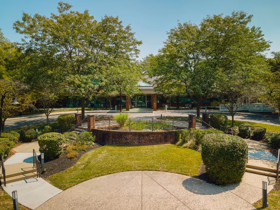 Front entrance of a facility with a landscaped circular brick planter, trees, and walkways leading to the building.