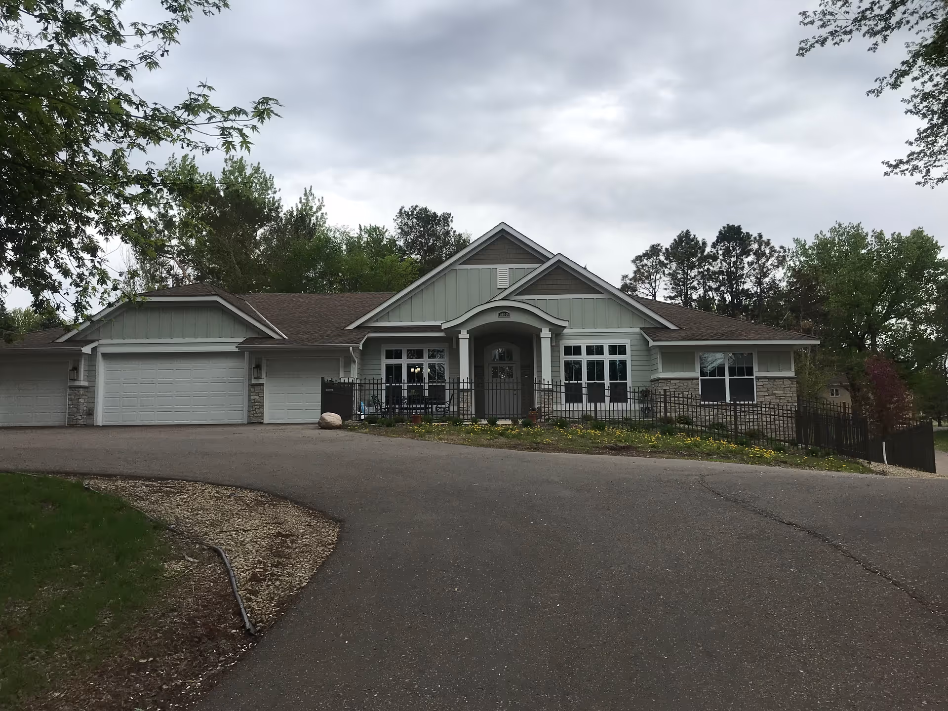 Front exterior view of a single-story residential building with a three-car garage, light gray siding, stone accents, and a covered entrance with columns. The driveway curves up to the entrance, and there are trees and greenery surrounding the property under a cloudy sky.