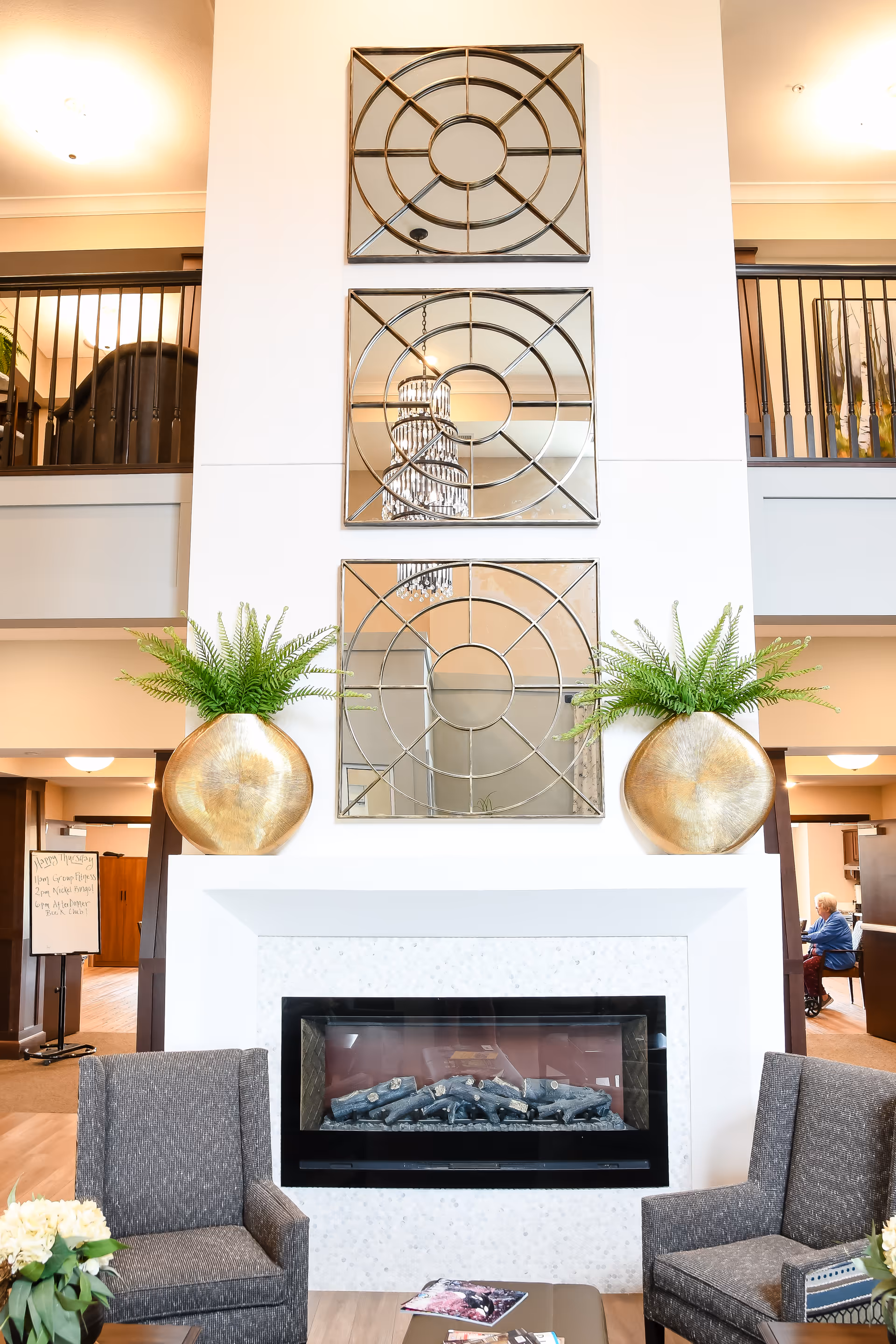 Lobby seating area with a modern fireplace, three decorative mirrors above it, large gold planters with ferns, and two upholstered chairs.