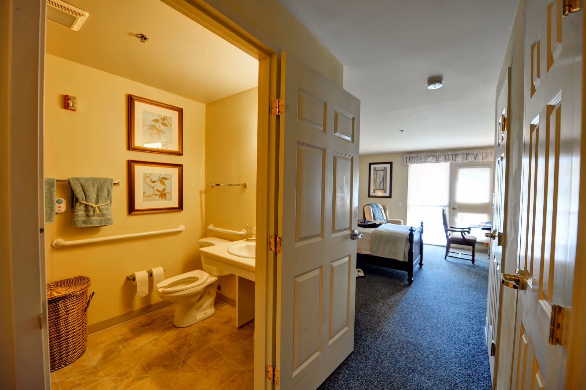 View of a senior living facility room showing an open door to a bathroom on the left with a toilet, sink, towel rack with towels, and framed artwork on the wall. The room beyond has a bed, chair, framed picture, and large windows with blinds letting in natural light.