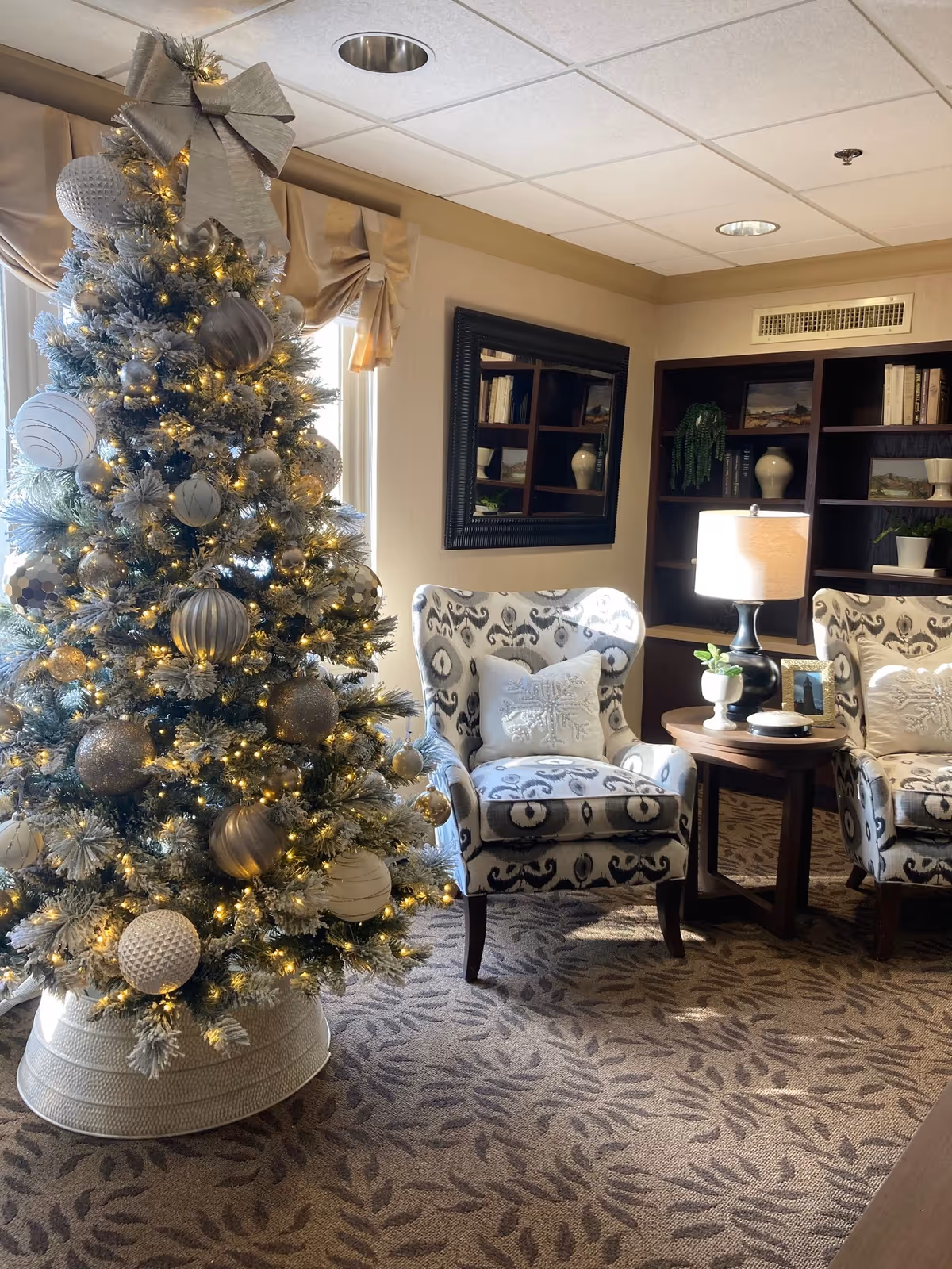A decorated Christmas tree beside two patterned armchairs and a side table with a lamp in a cozy sitting area.