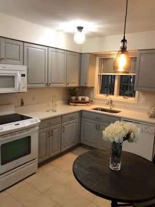 A modern kitchen with gray cabinets, a white microwave above a white stove, a white dishwasher, and a sink under a window. There is a round dark wooden table in the foreground with a glass vase holding white flowers. The kitchen is well-lit with ceiling and hanging lights.