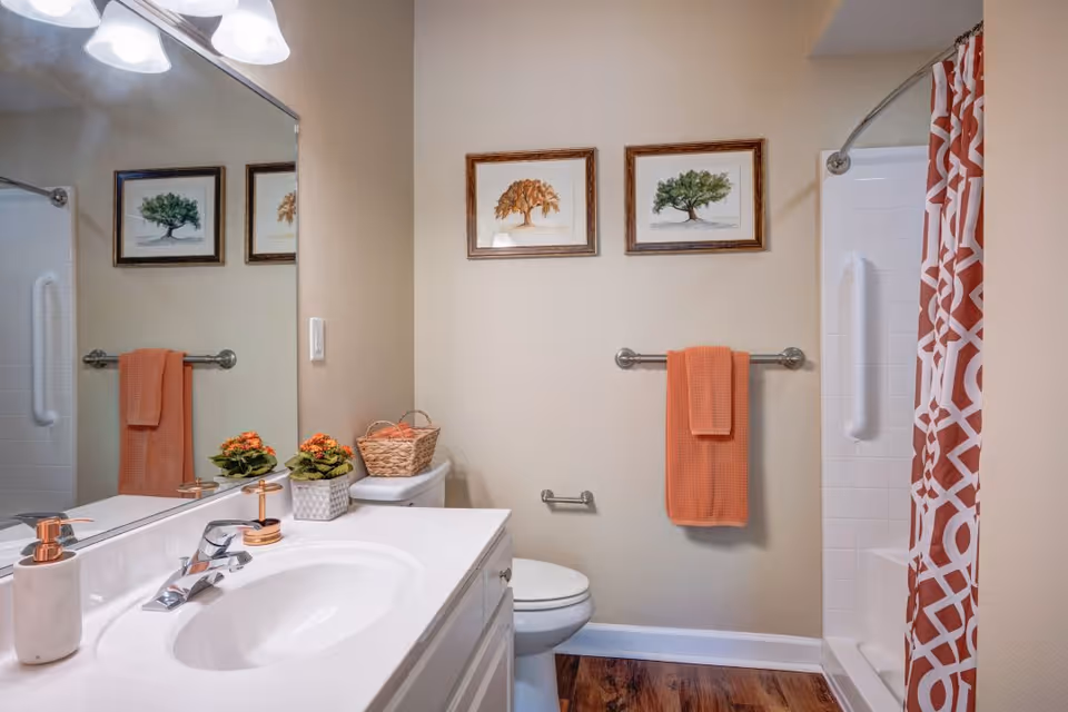 A clean and well-lit bathroom featuring a white sink with a chrome faucet, a soap dispenser, and two small potted plants on the countertop. Above the sink is a large mirror reflecting part of the room. On the wall, there are two framed pictures of trees and an orange towel hanging on a towel rack. To the right, there is a white toilet and a shower with a white tiled wall and an orange patterned shower curtain. The floor has a wood-like finish.