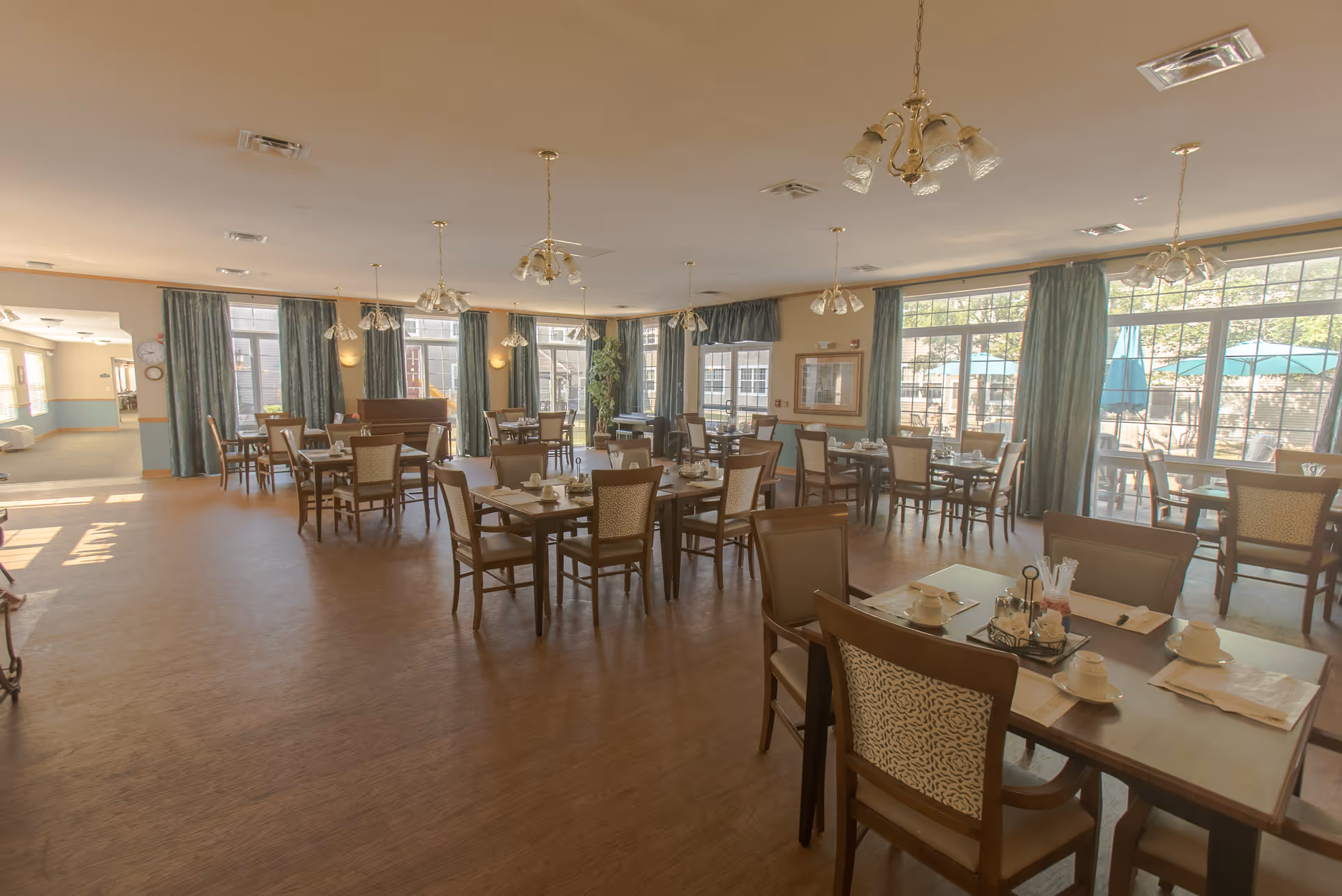 A spacious dining room with multiple wooden tables and chairs arranged neatly. Each table is set with white cups, saucers, napkins, and condiments. Large windows with green curtains allow natural light to fill the room, and there are several hanging light fixtures on the ceiling. Outside the windows, patio umbrellas are visible.